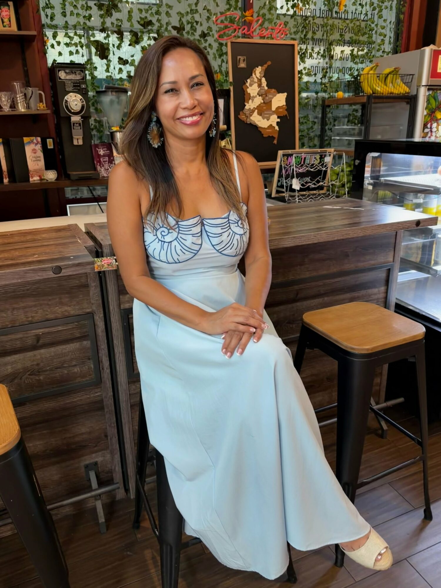 Woman in a light blue dress sitting on a barstool in a cafe, smiling, with a wooden counter and shelves behind her.