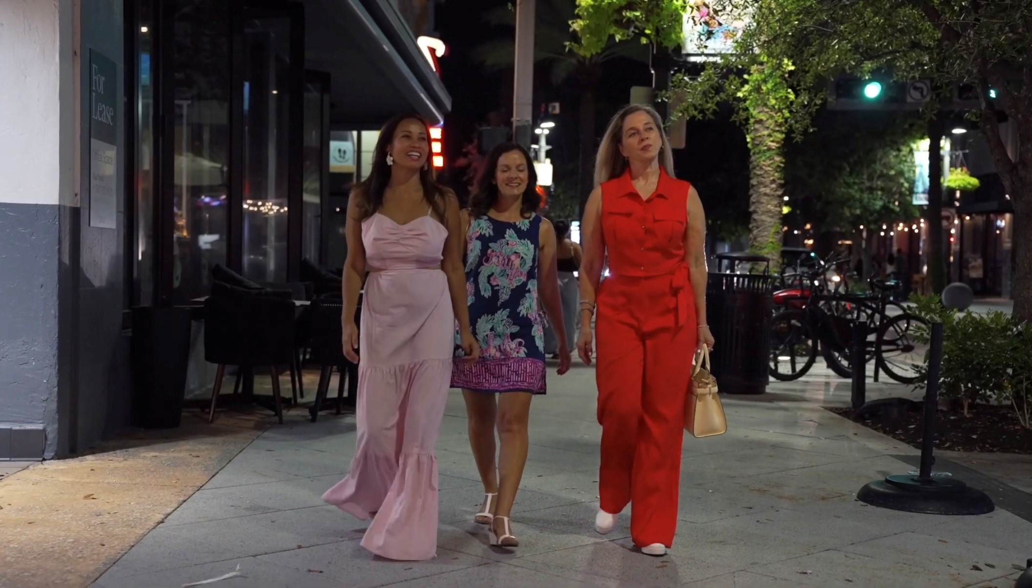 Three women walking on a city sidewalk at night, illuminated by streetlights and storefronts, with trees and bicycles nearby.