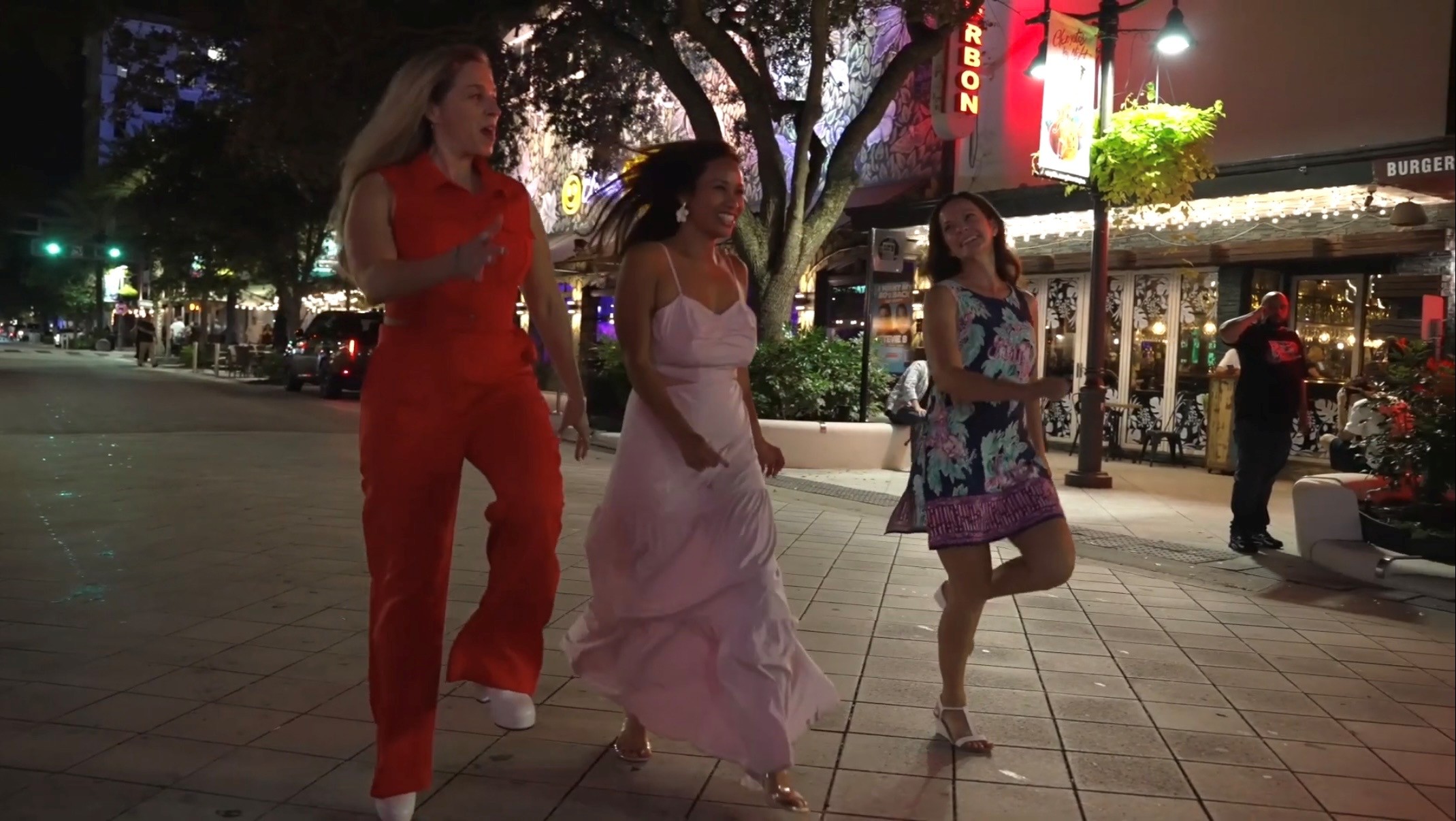 Three women walking on a city sidewalk at night, illuminated by streetlights and shop signs.