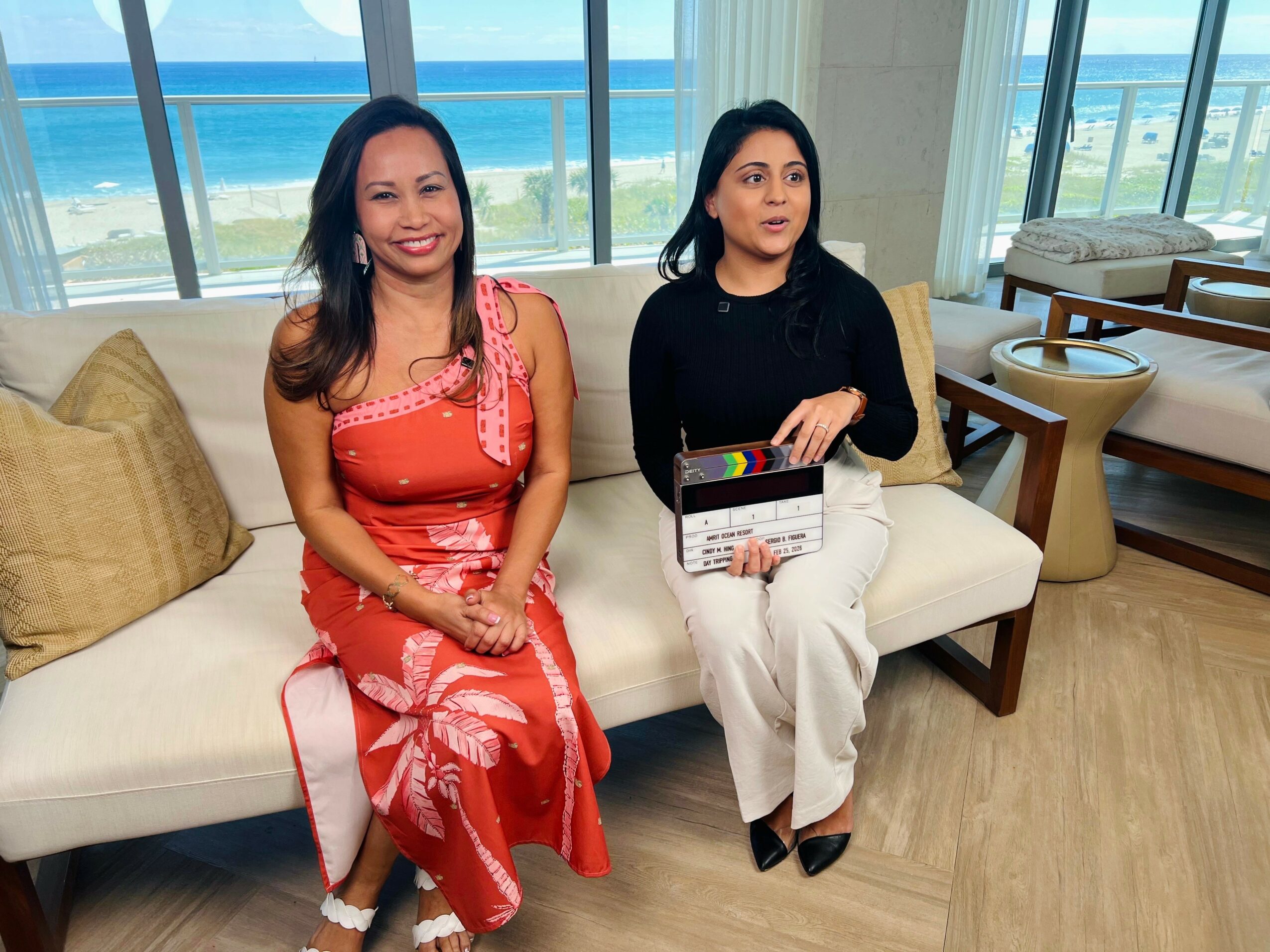 Two women sitting on a white sofa in a bright room with large windows showing a beach view, one smiling, one holding a clapperboard.