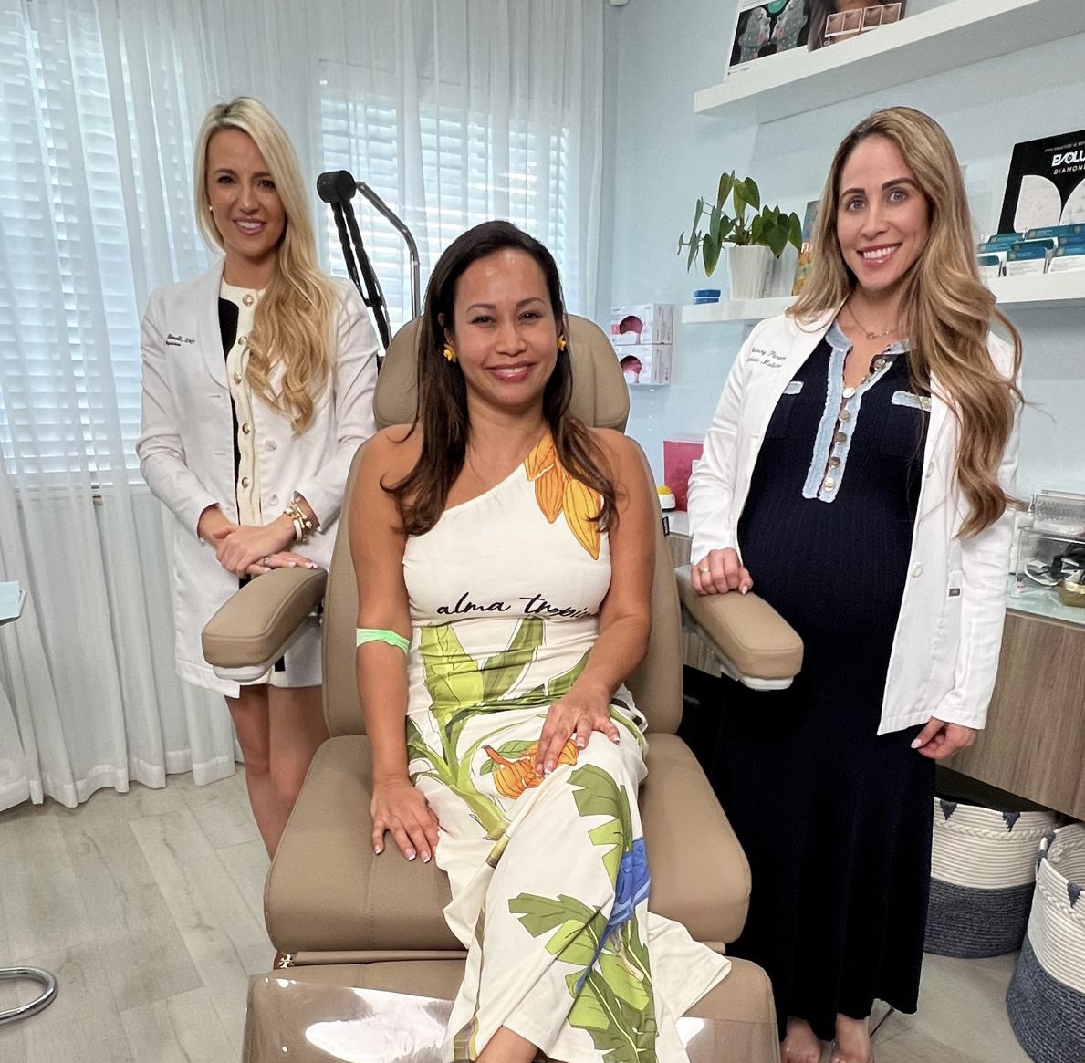 Three women in a medical office, one seated and two standing, smiling, with medical equipment and shelves behind them.