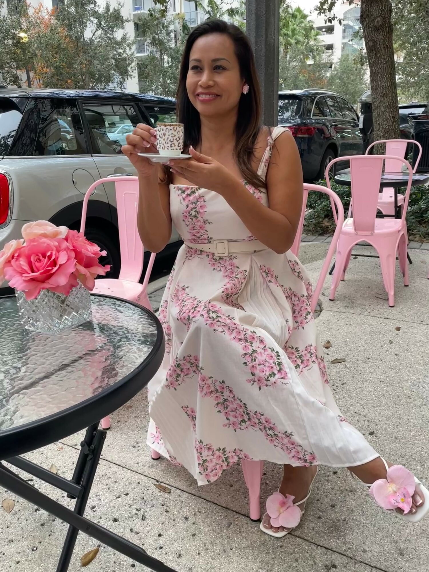 Woman in white floral dress sitting outdoors, holding a cup, pink flowers on table, pink chairs, trees, parked cars in background.