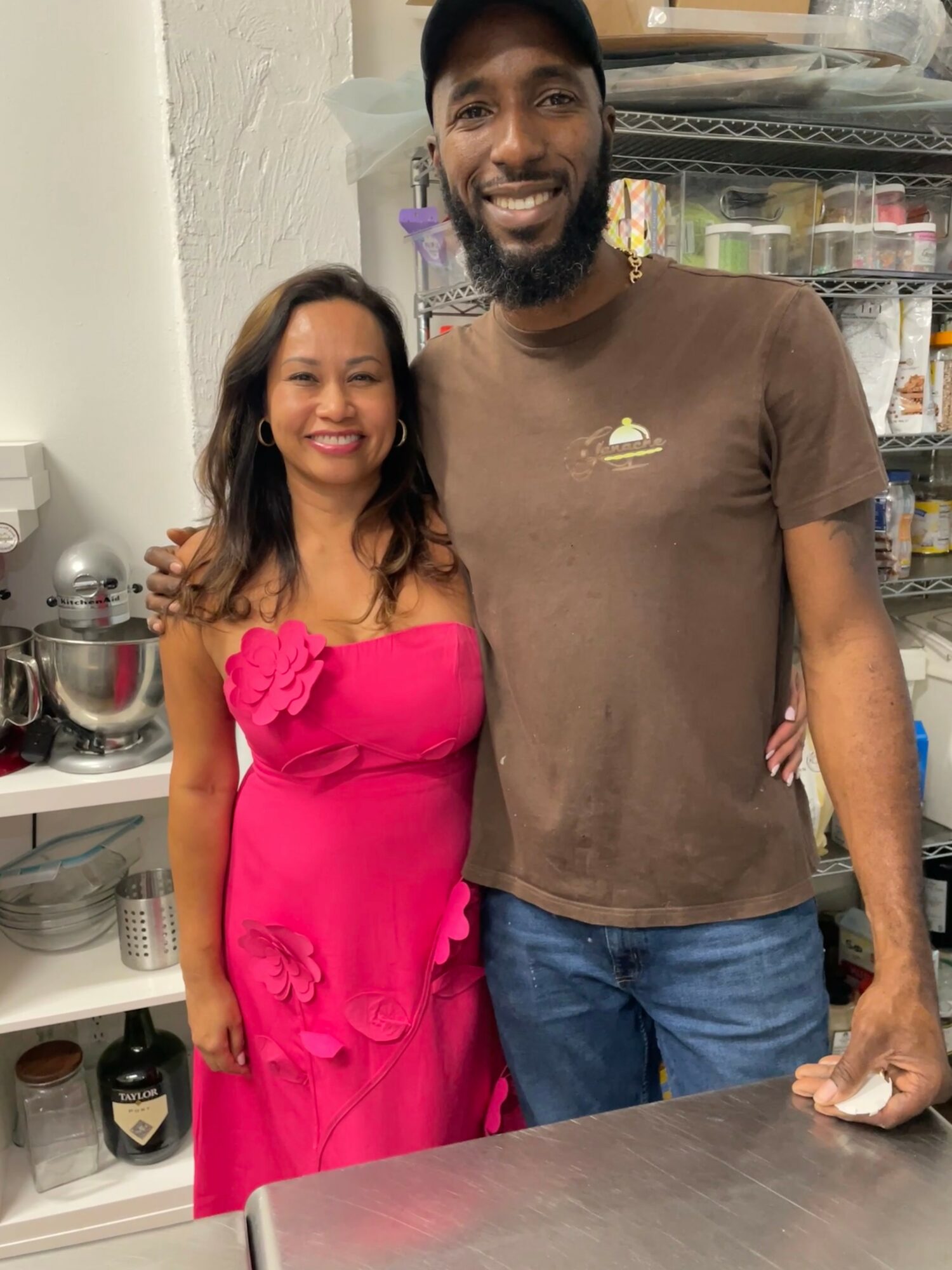 A woman in a pink dress and a man in a brown t-shirt stand together in a kitchen, smiling.