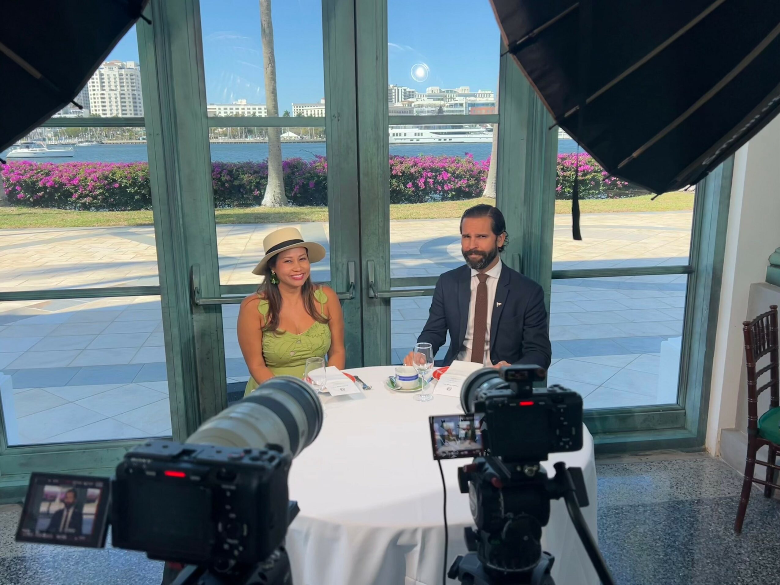 Two people sit at a table with cameras recording them, large glass windows with outdoor scenery behind, sunny day.
