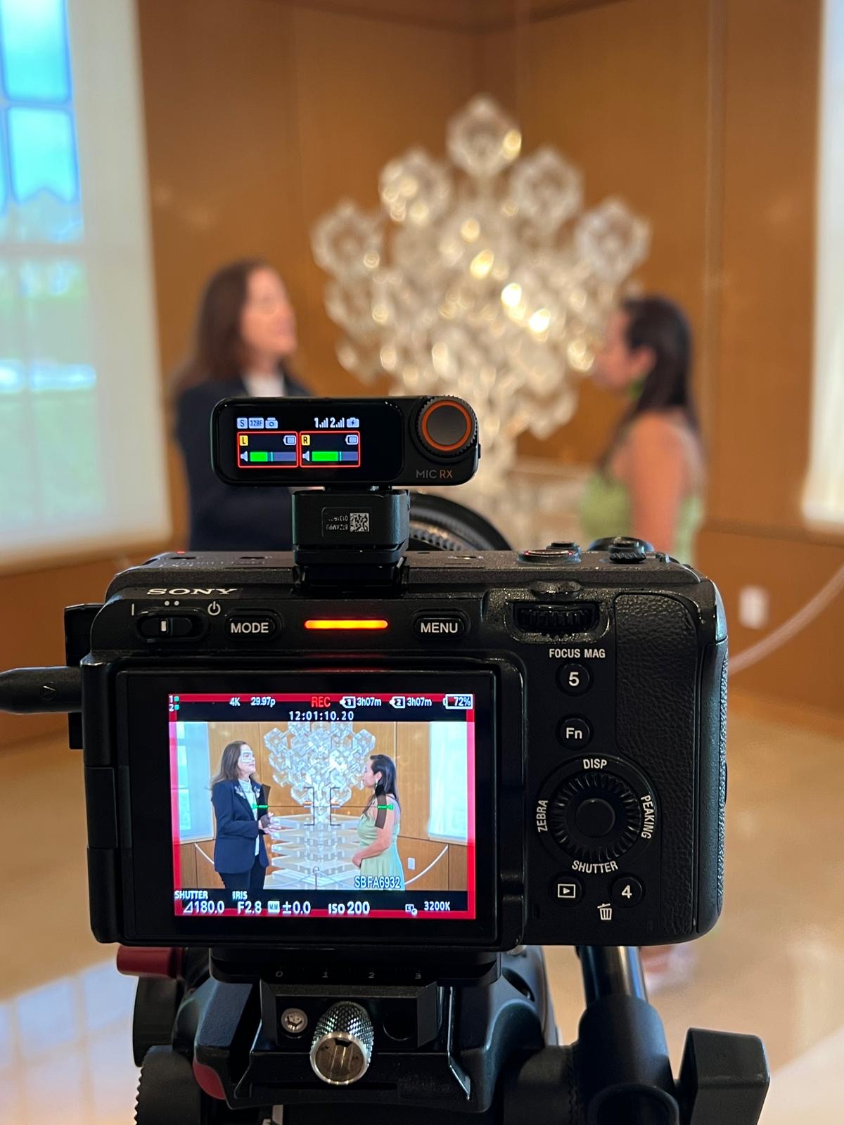 Camera viewfinder shows two women standing in front of a decorative wall with a large, illuminated art piece.