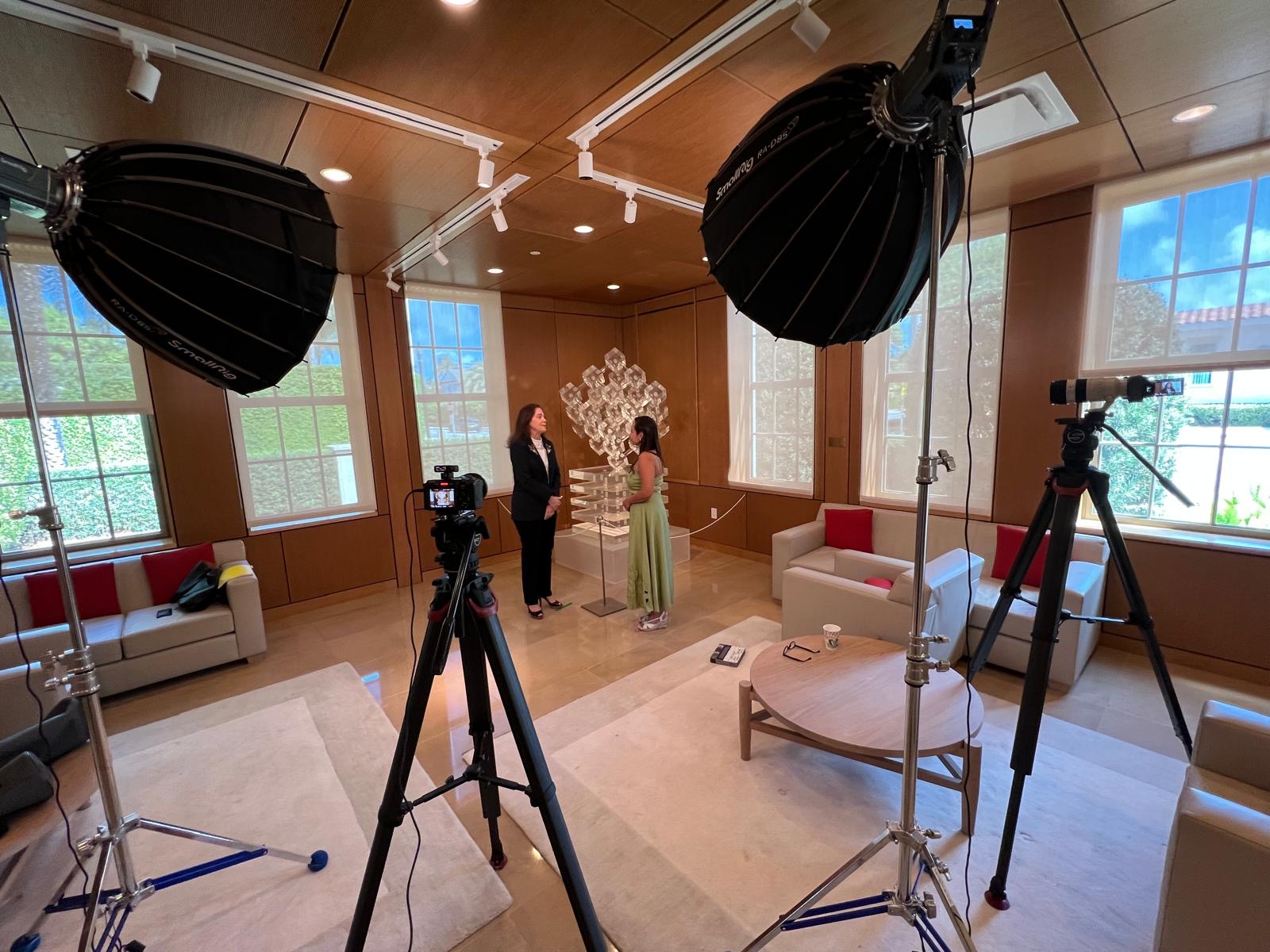 Two people stand in a room with large windows, surrounded by photography equipment and studio lights, with a chandelier and furniture.
