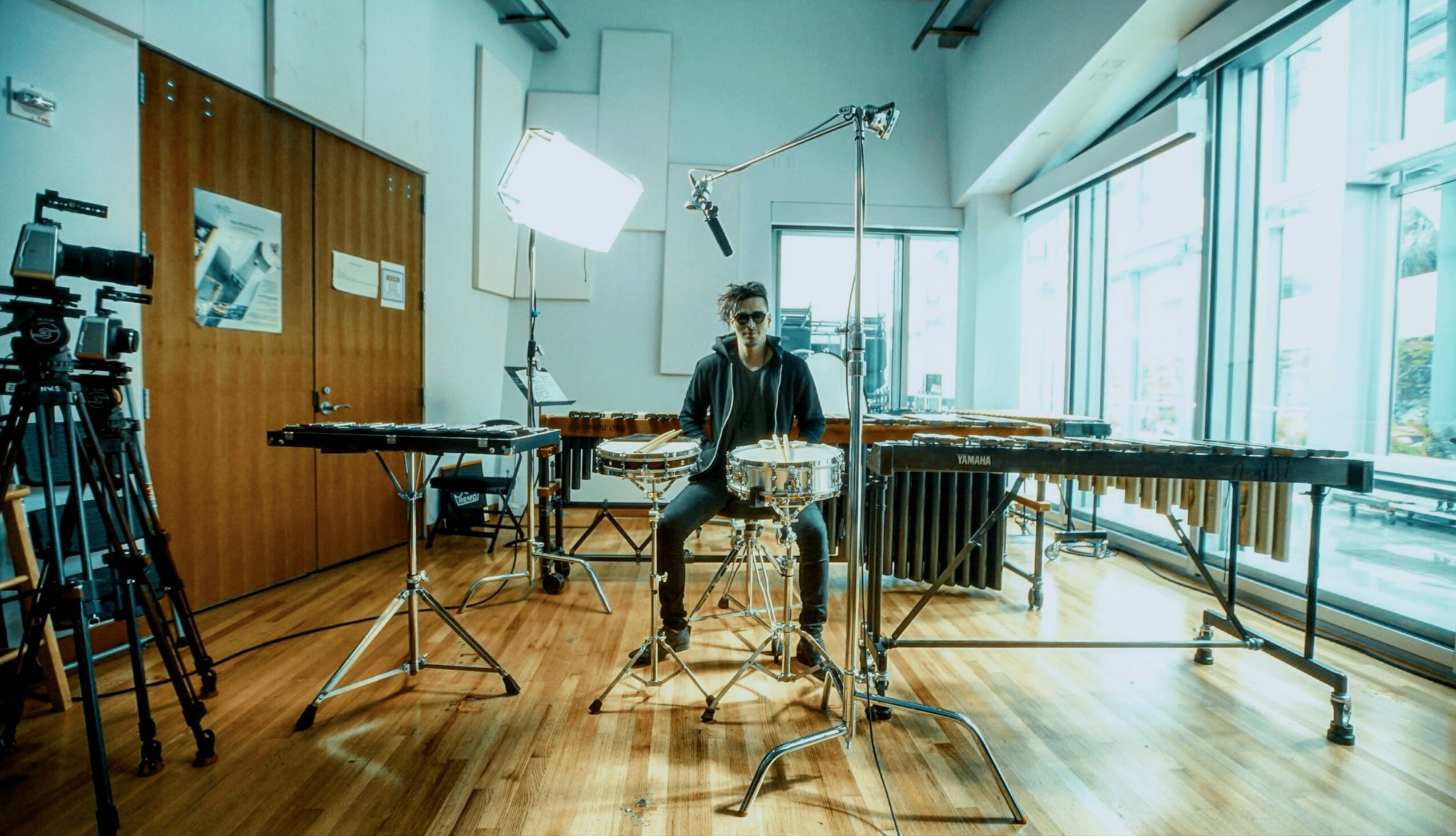 Person sitting at a table with musical instruments in a bright studio with large windows, filming equipment around.