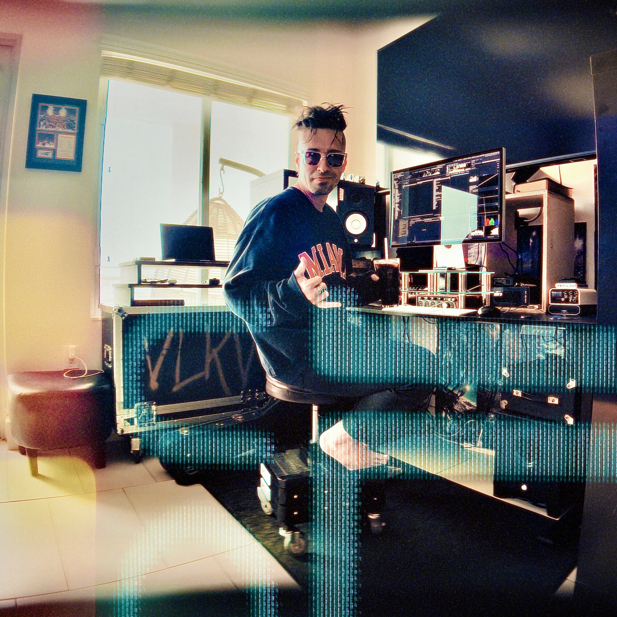 Person with sunglasses sitting at a desk with multiple computer monitors and equipment, waving at the camera, in a bright room.