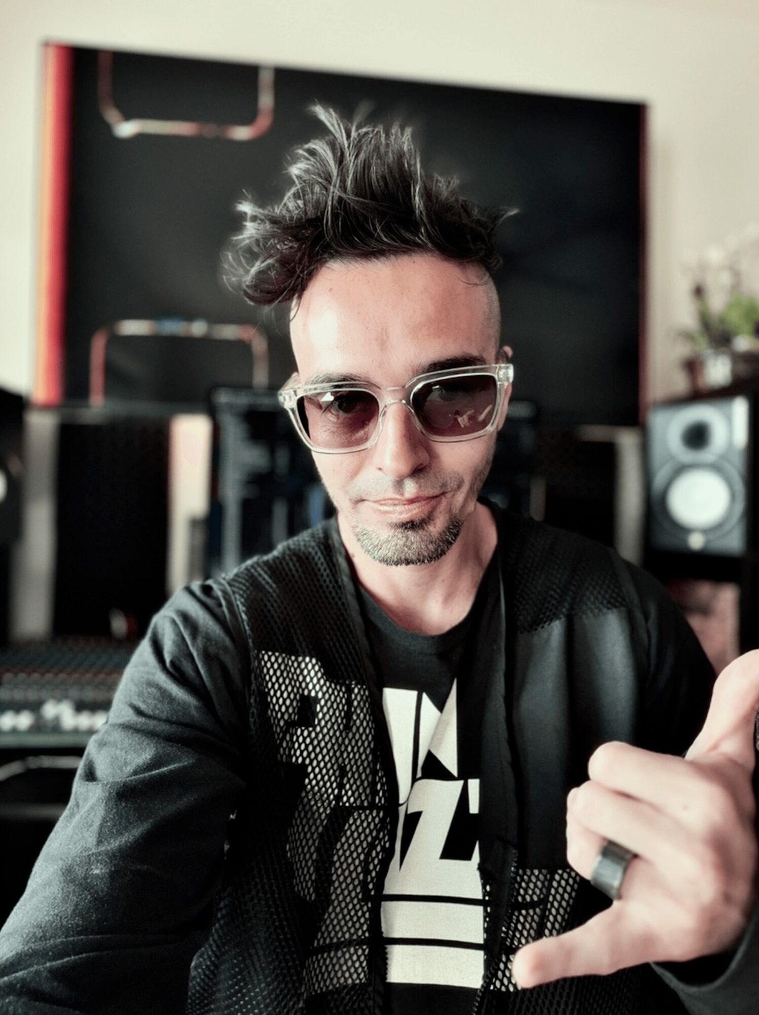 Person with spiky hair wearing sunglasses and a black jacket, making a hand gesture, in a music studio.