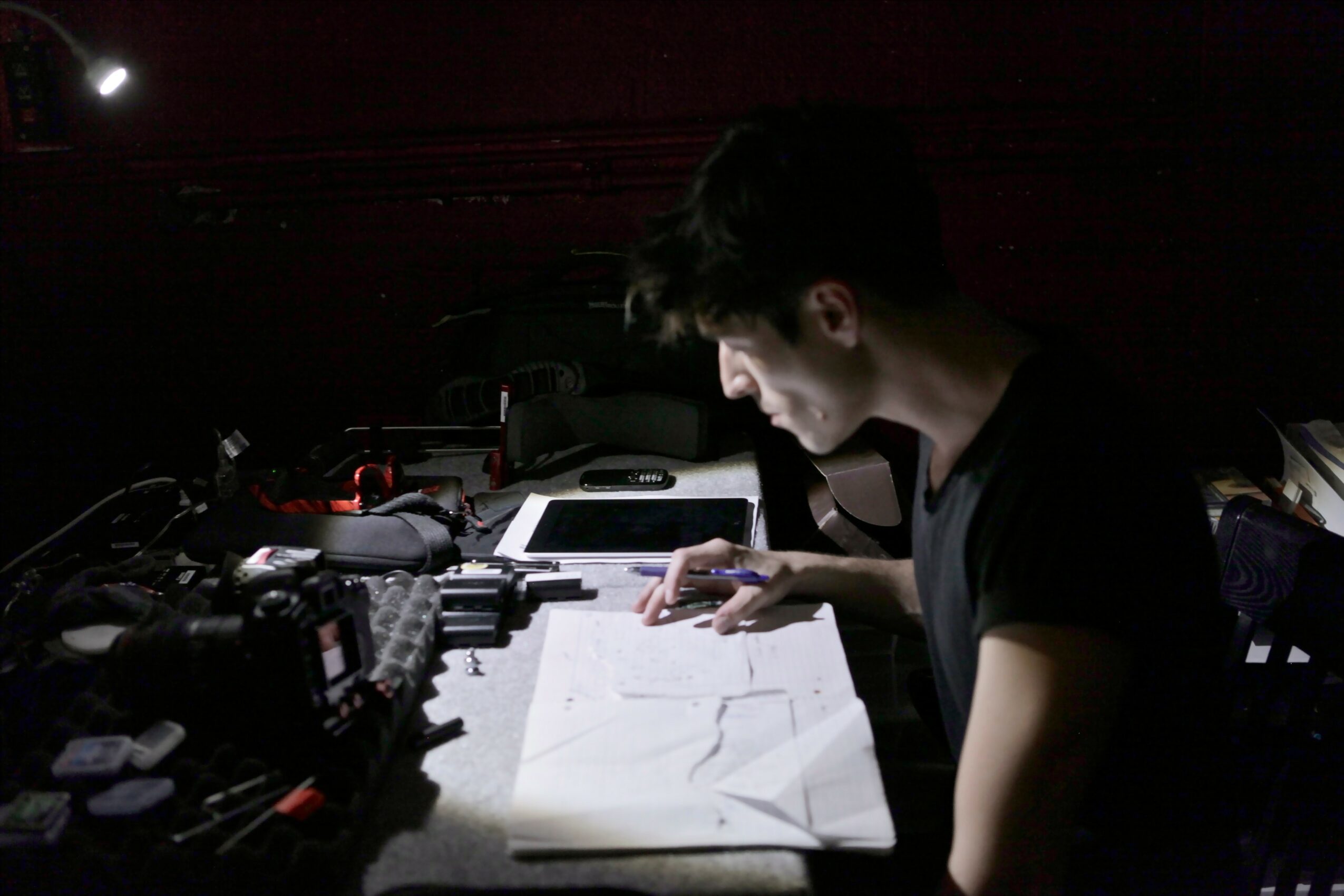 Young person studying with papers and electronic devices on a dark desk.