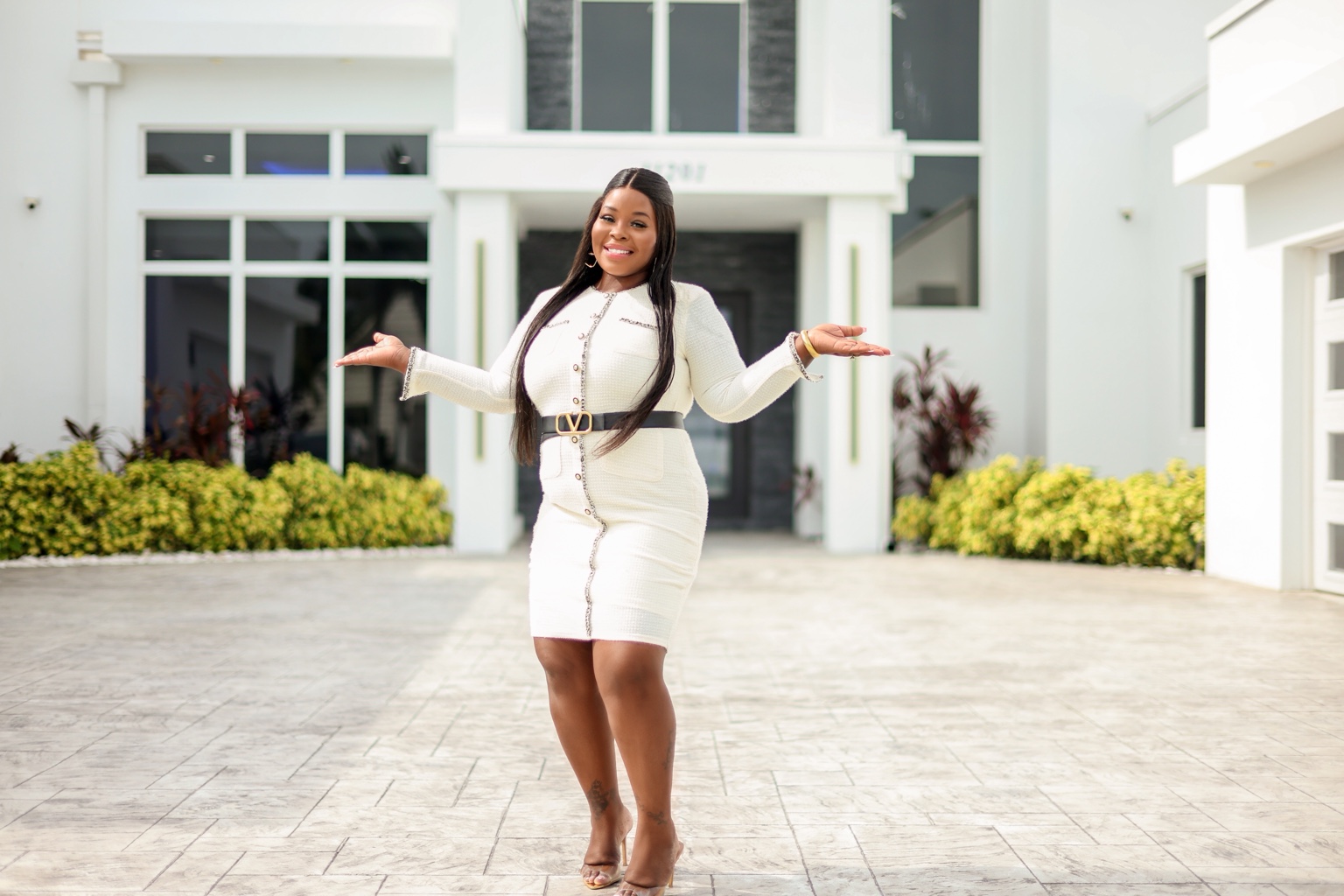 Smiling woman in white dress with arms outstretched standing in front of a modern white building with greenery