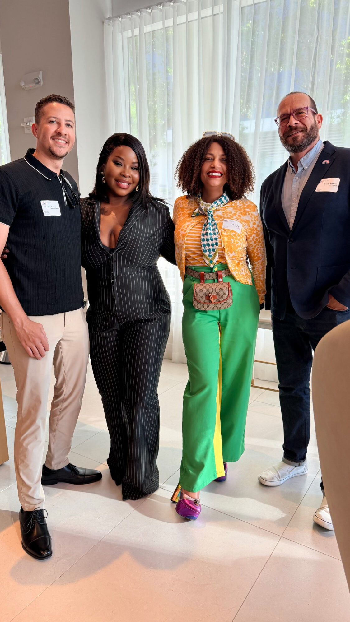Four people standing indoors near a window, smiling, dressed in business casual and colorful attire.