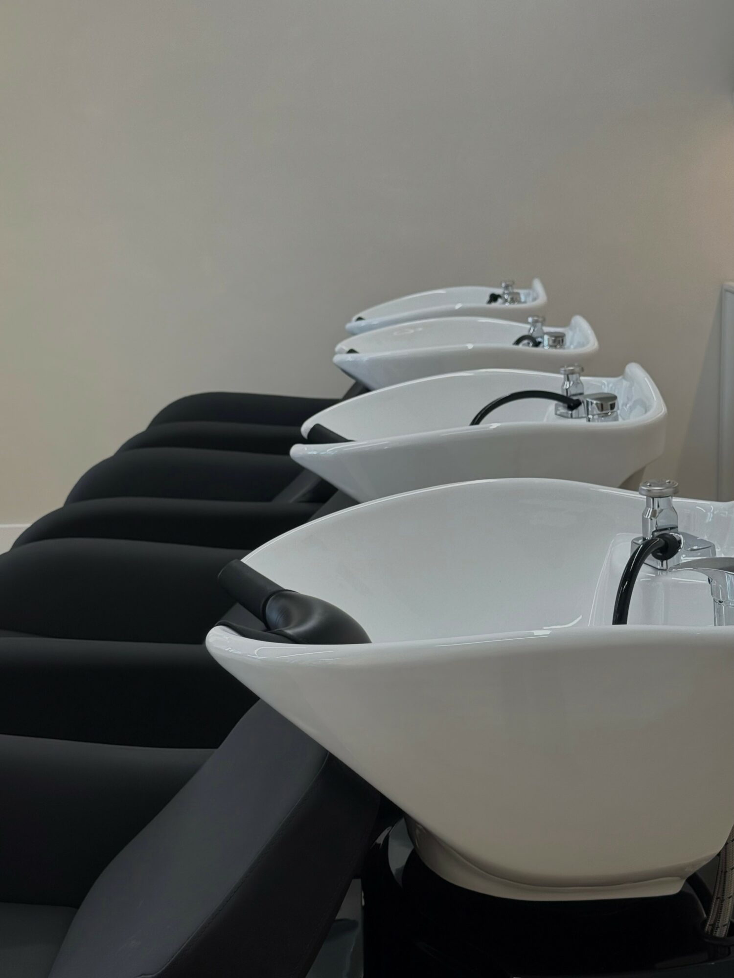 Four white salon washbasins with black chairs in a row against a plain wall.