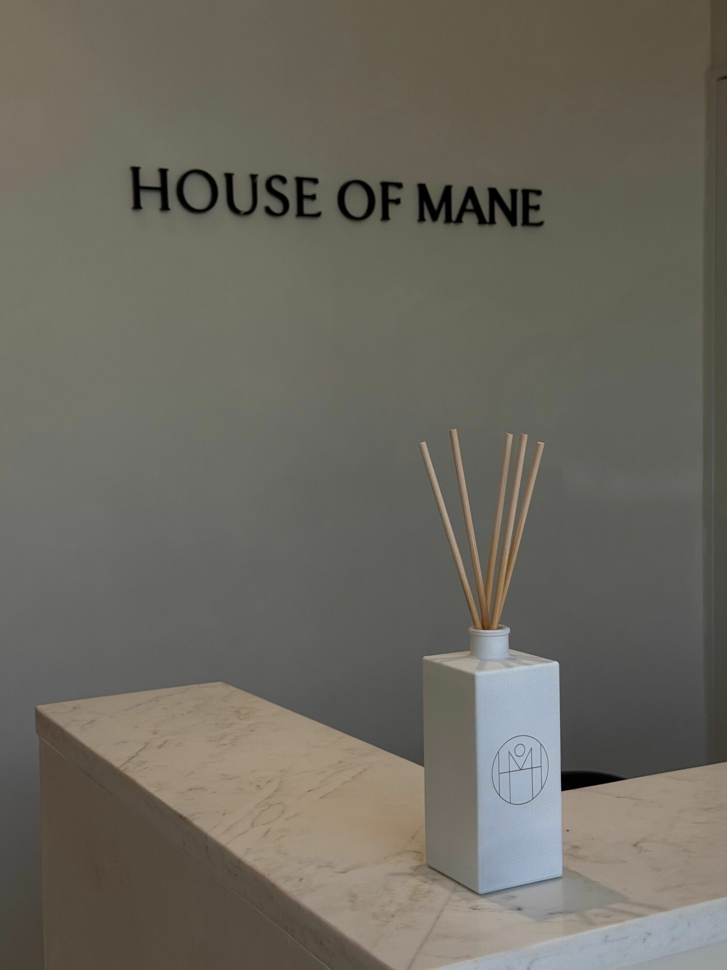 A white diffuser with wooden sticks on a light-colored surface, with text 'HOUSE OF MANE' on a gray wall.