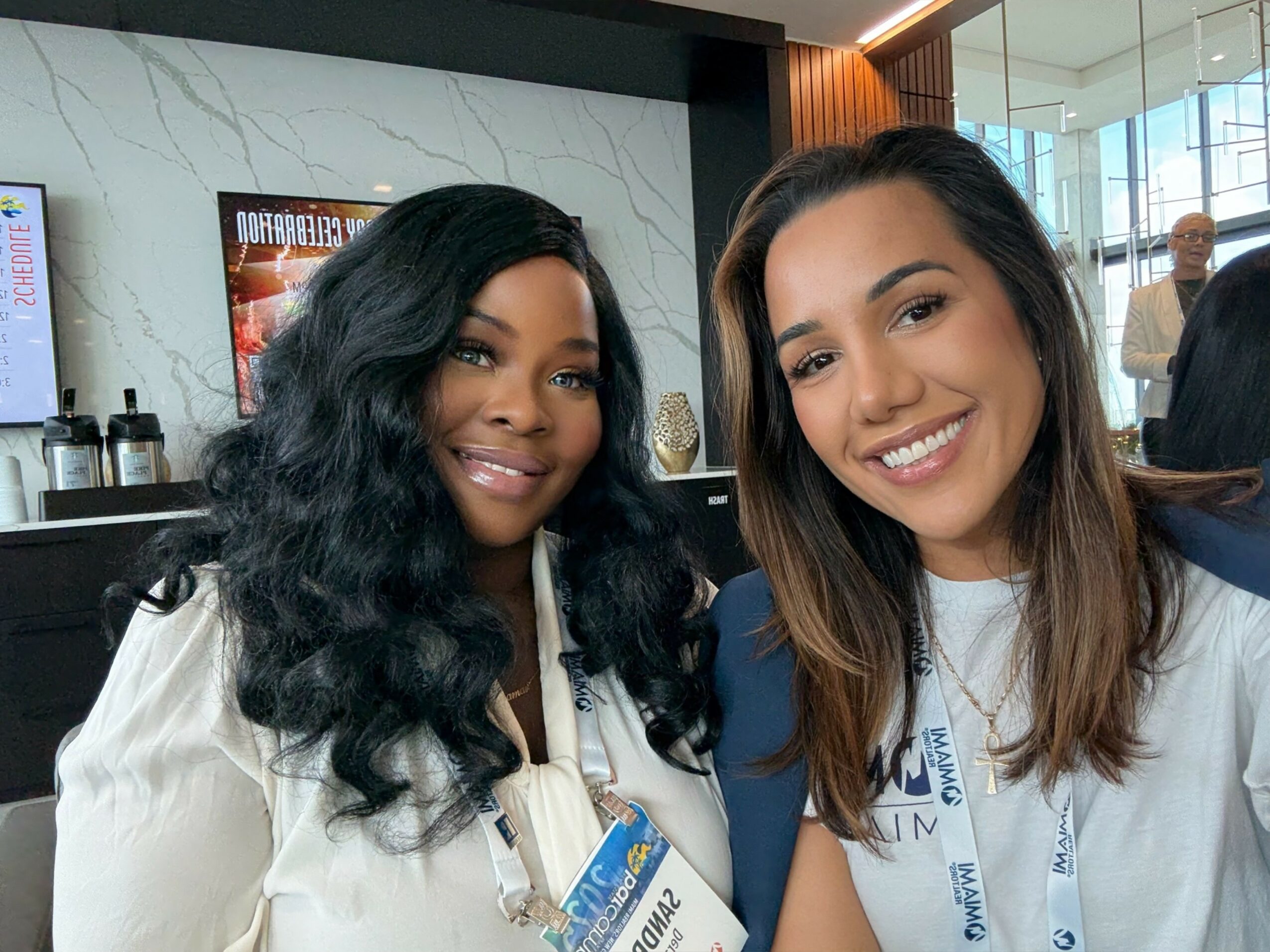 Two women smiling and posing for a photo indoors, wearing white lanyards, with a modern background.