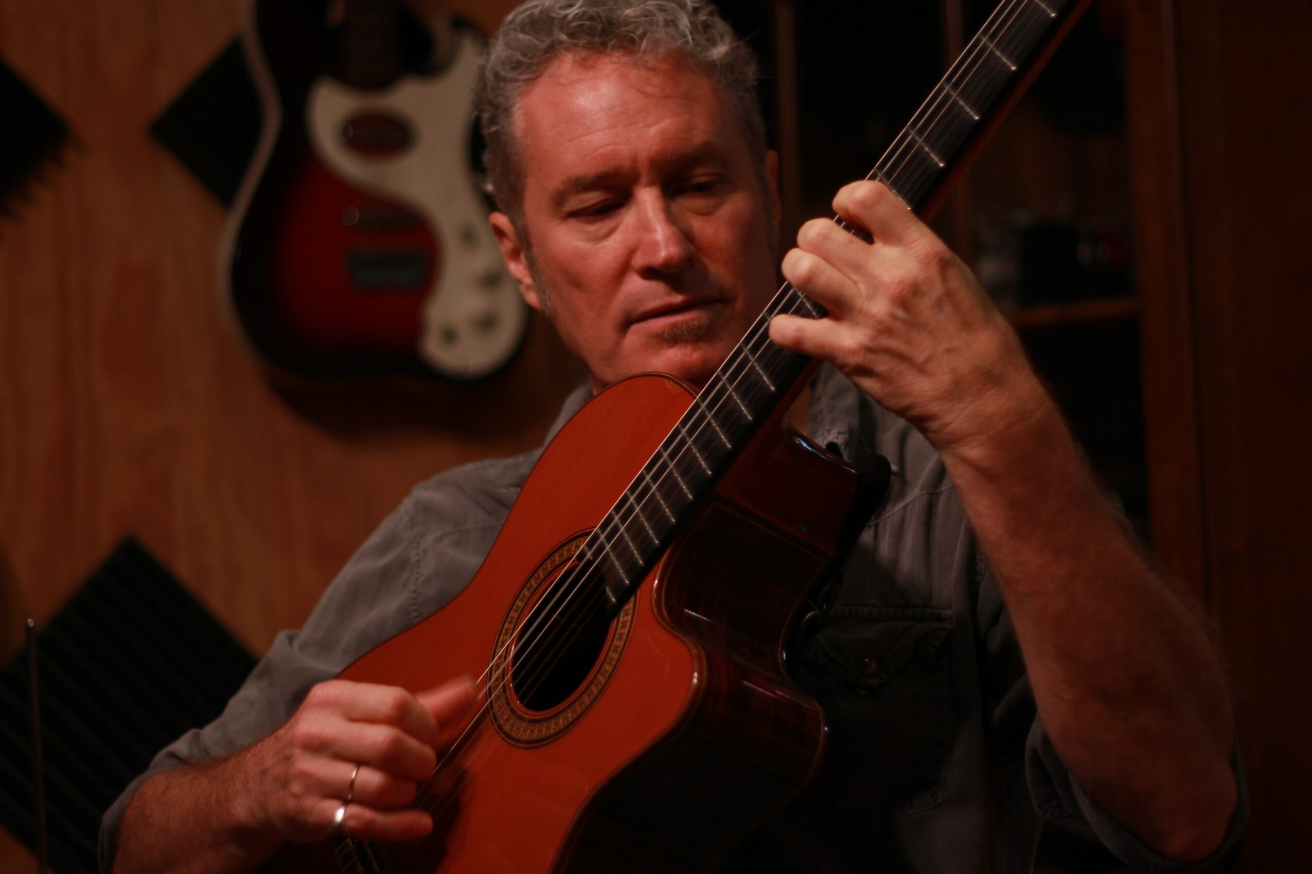 Man playing an acoustic guitar indoors, with guitars hanging on the wall behind him.