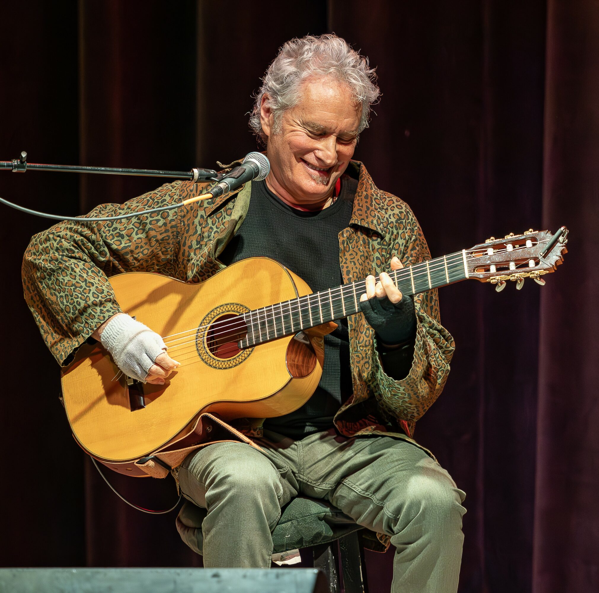 Person with gray curly hair playing an acoustic guitar and singing into a microphone on stage.