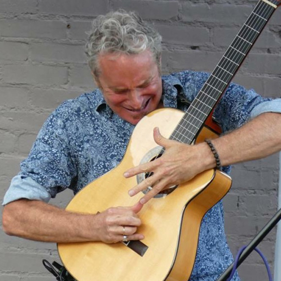 Elderly man with curly gray hair playing an acoustic guitar passionately, eyes closed, against a gray brick wall.