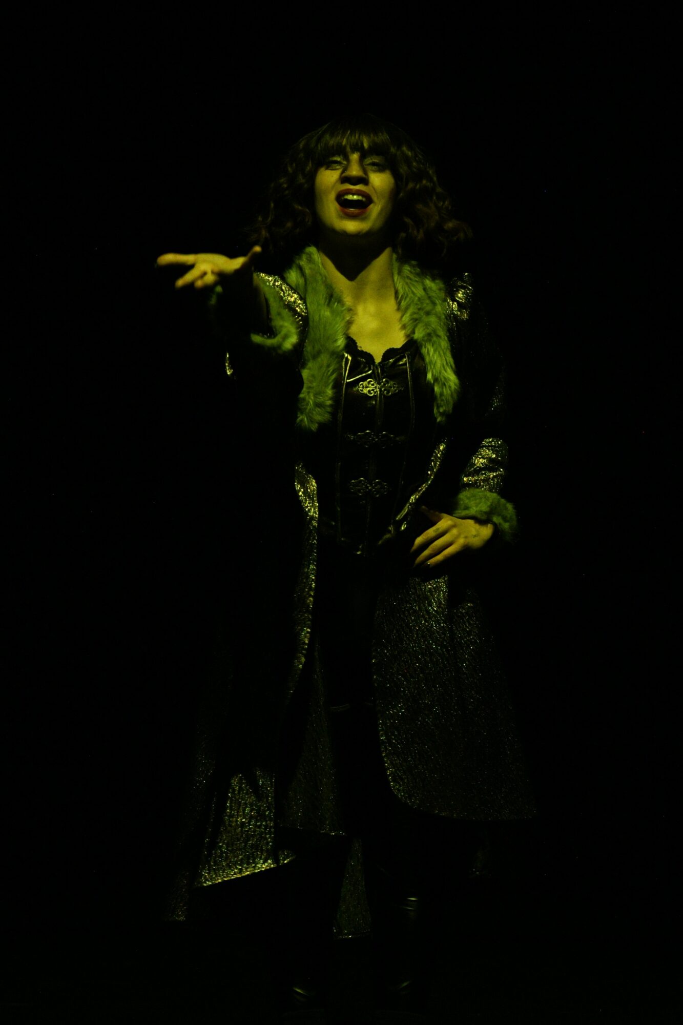Person with curly hair singing or speaking, wearing dark clothing and a patterned coat, with a dark background.