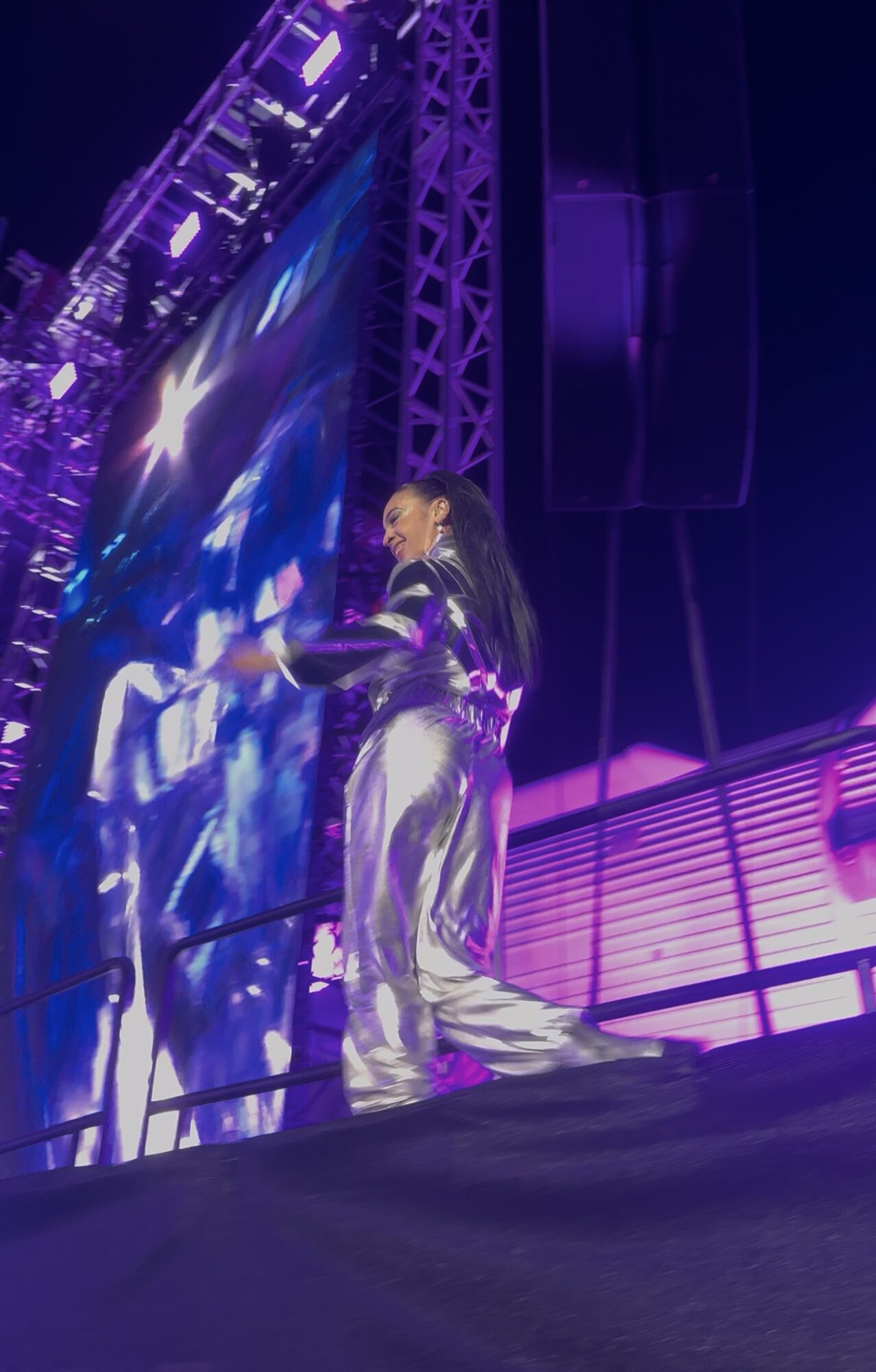 Performer on stage with purple lighting, wearing shiny silver outfit, facing a large screen, with stage equipment visible.