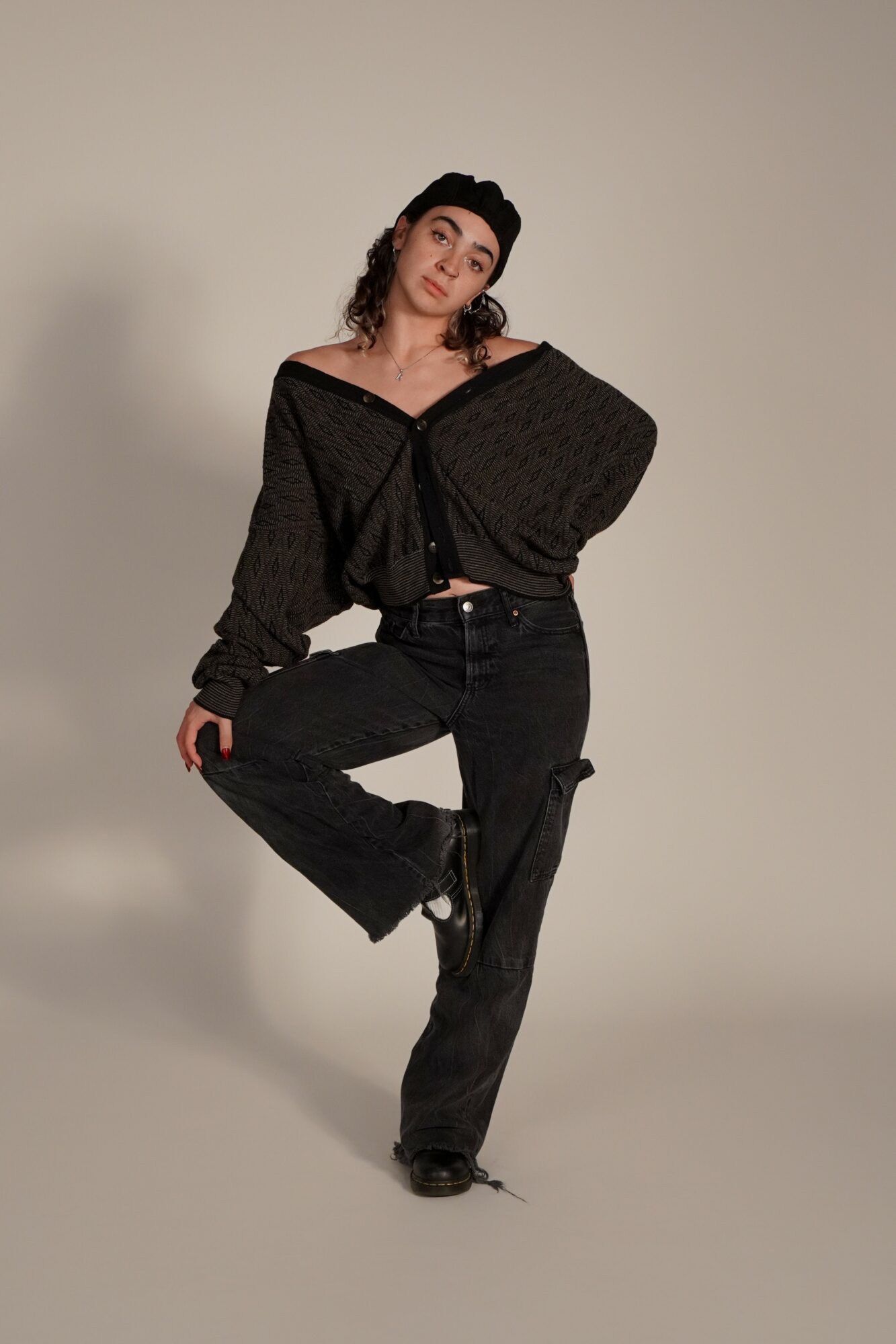 Young woman with curly hair in a black outfit, standing with one leg raised, against a plain background.
