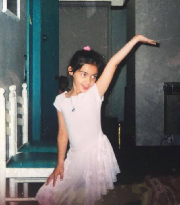 Young girl in a white dress with a pink hair accessory, posing with one arm raised in a room.