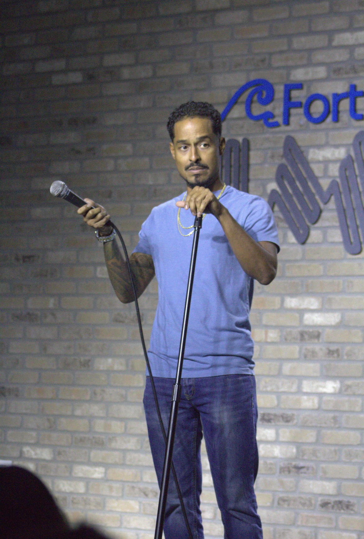 Man standing on stage holding a microphone, wearing a blue t-shirt and jeans, with a brick wall background and a blue logo.