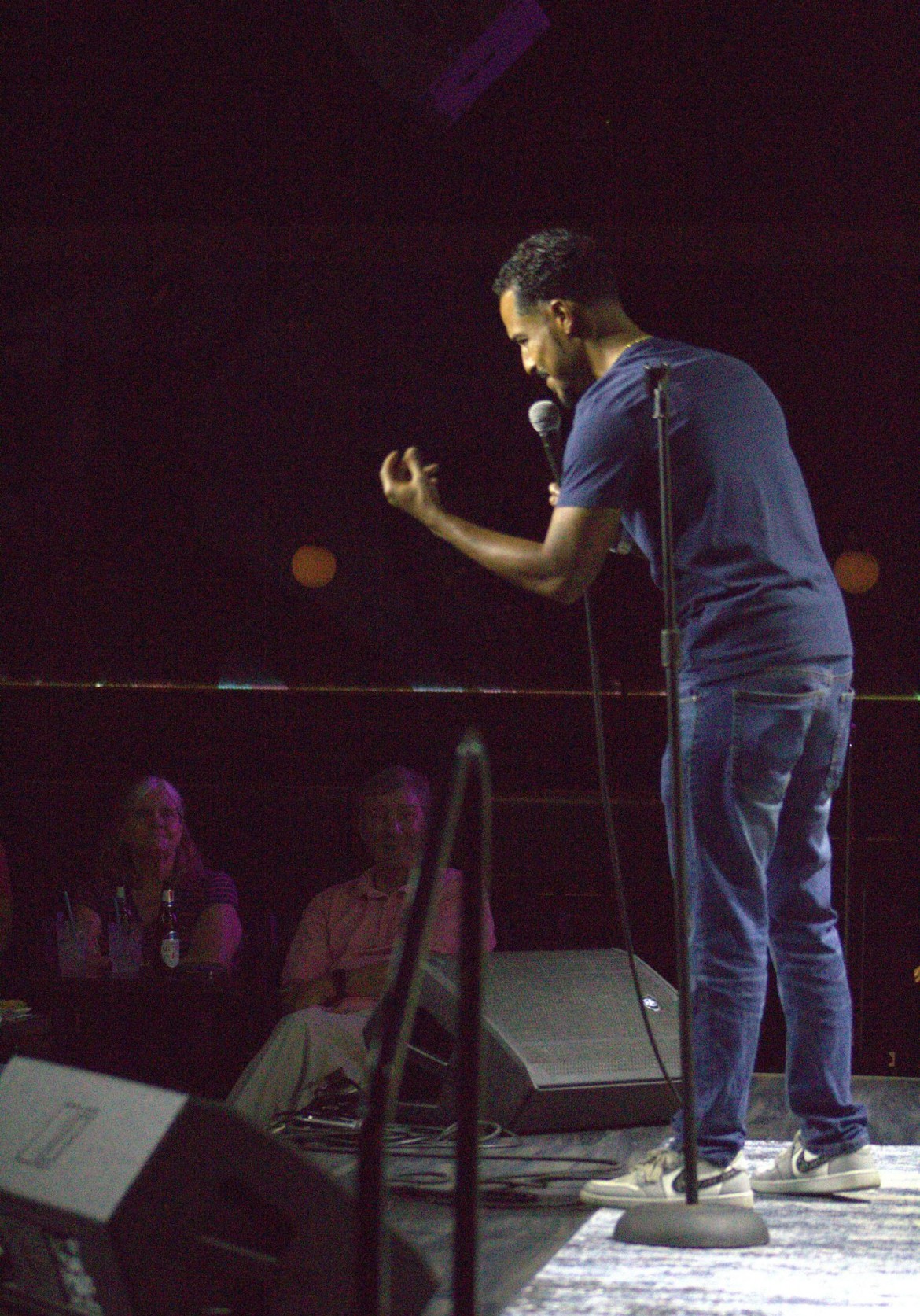 A man stands on stage, speaking into a microphone, with an audience watching in the background.