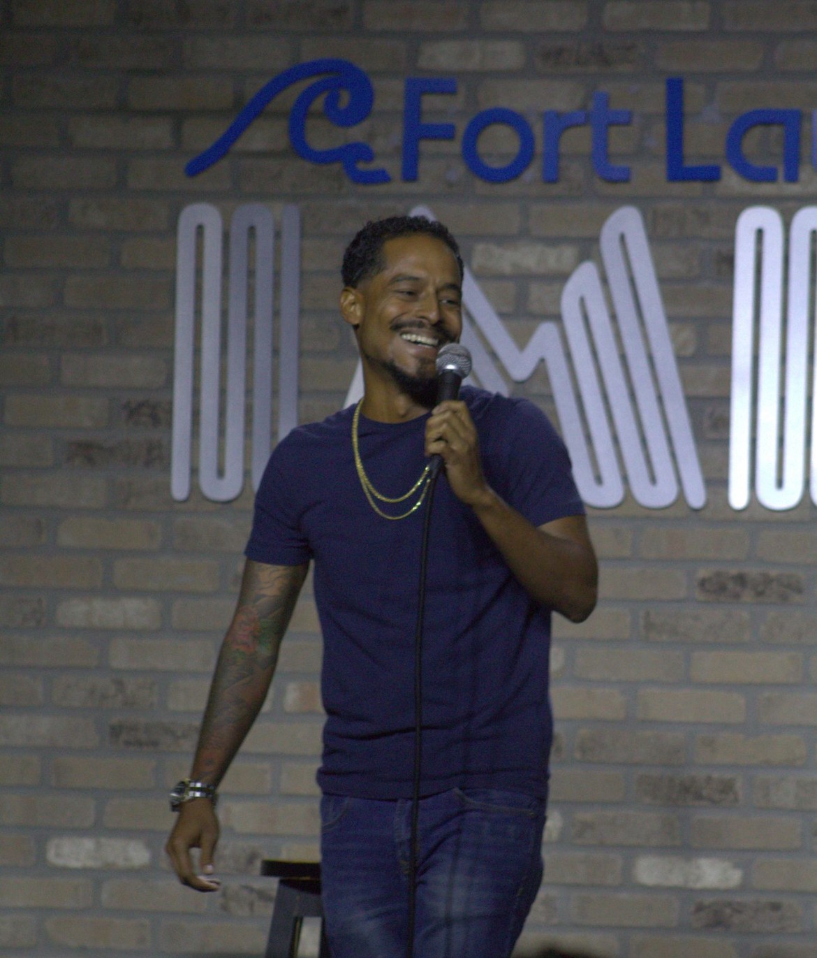 Man smiling and holding a microphone on stage with a brick wall background and partial sign reading 'Fort La'.