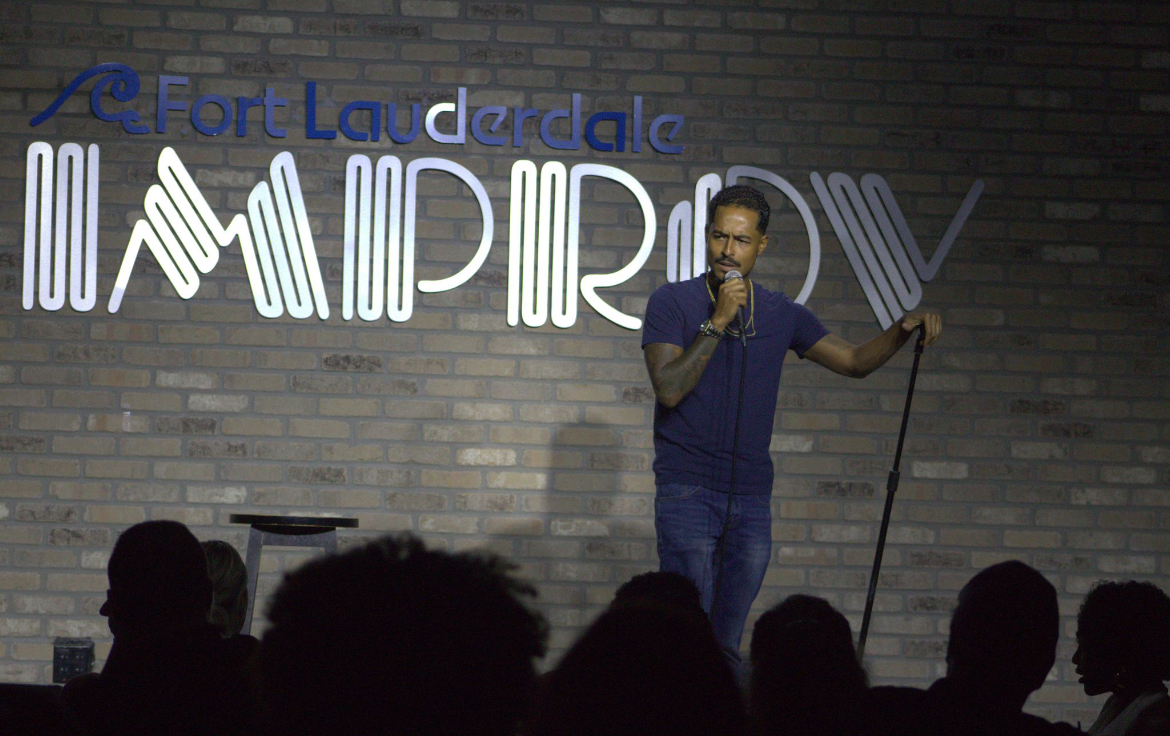 Man standing on stage with microphone and cane, audience silhouettes in foreground, brick wall background, illuminated sign behind him.
