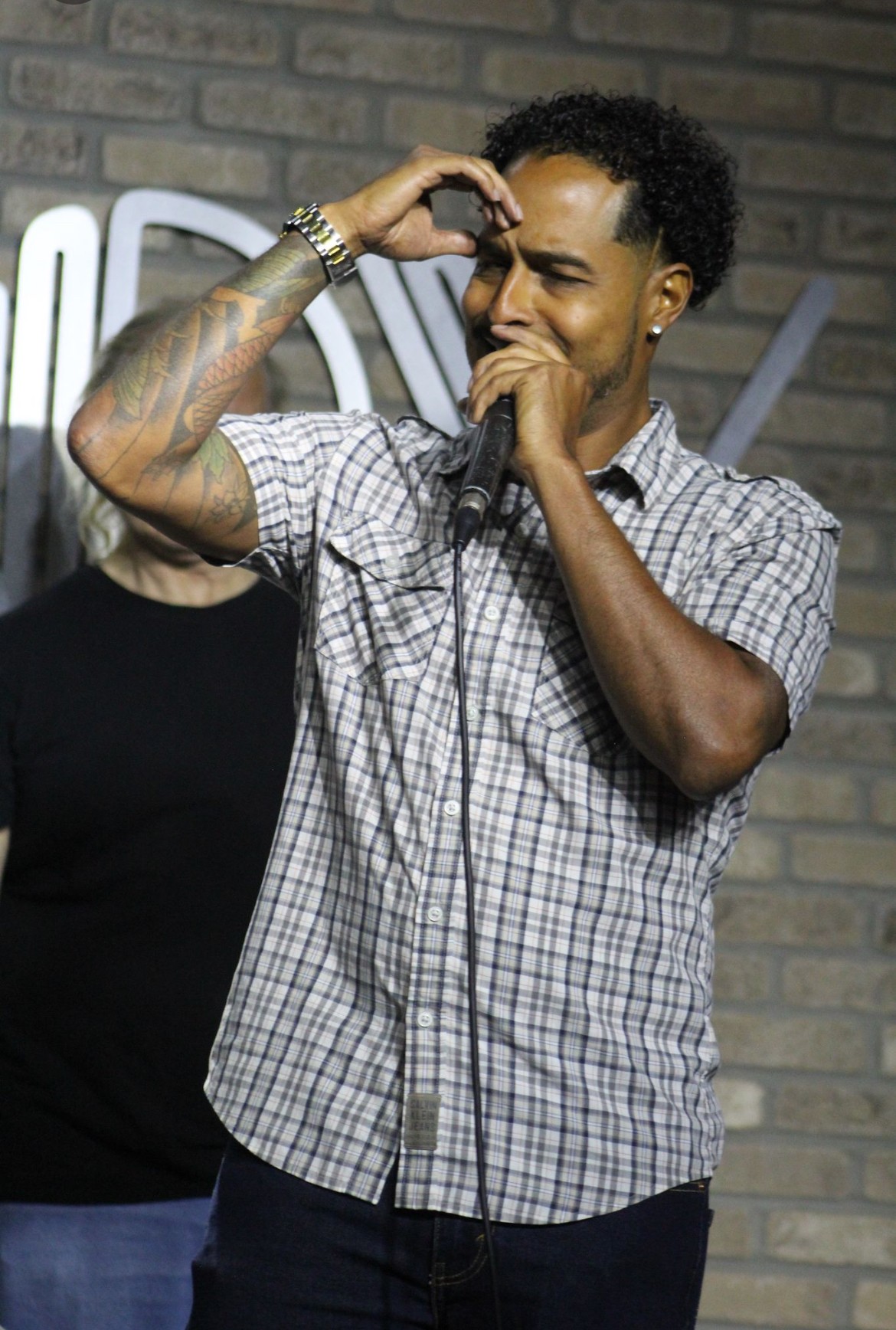 Young man with curly hair and tattoos, smiling and covering his mouth, standing in front of a brick wall.