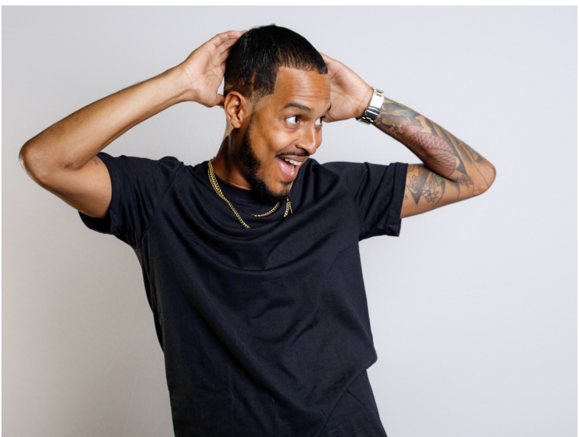 Person with short hair and tattoos, wearing a black t-shirt, smiling with hands on head, against a plain background.