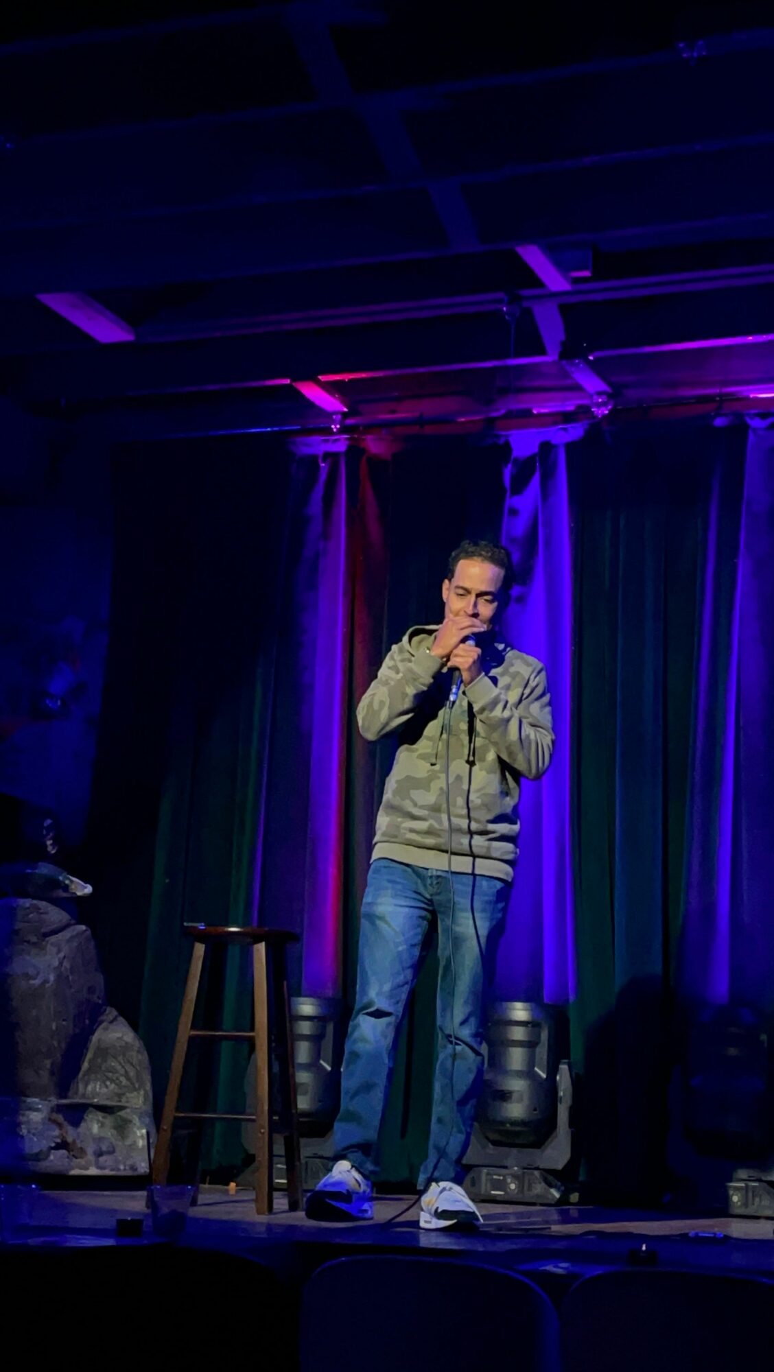 Person standing on stage with microphone, colorful lighting, and curtains in background.