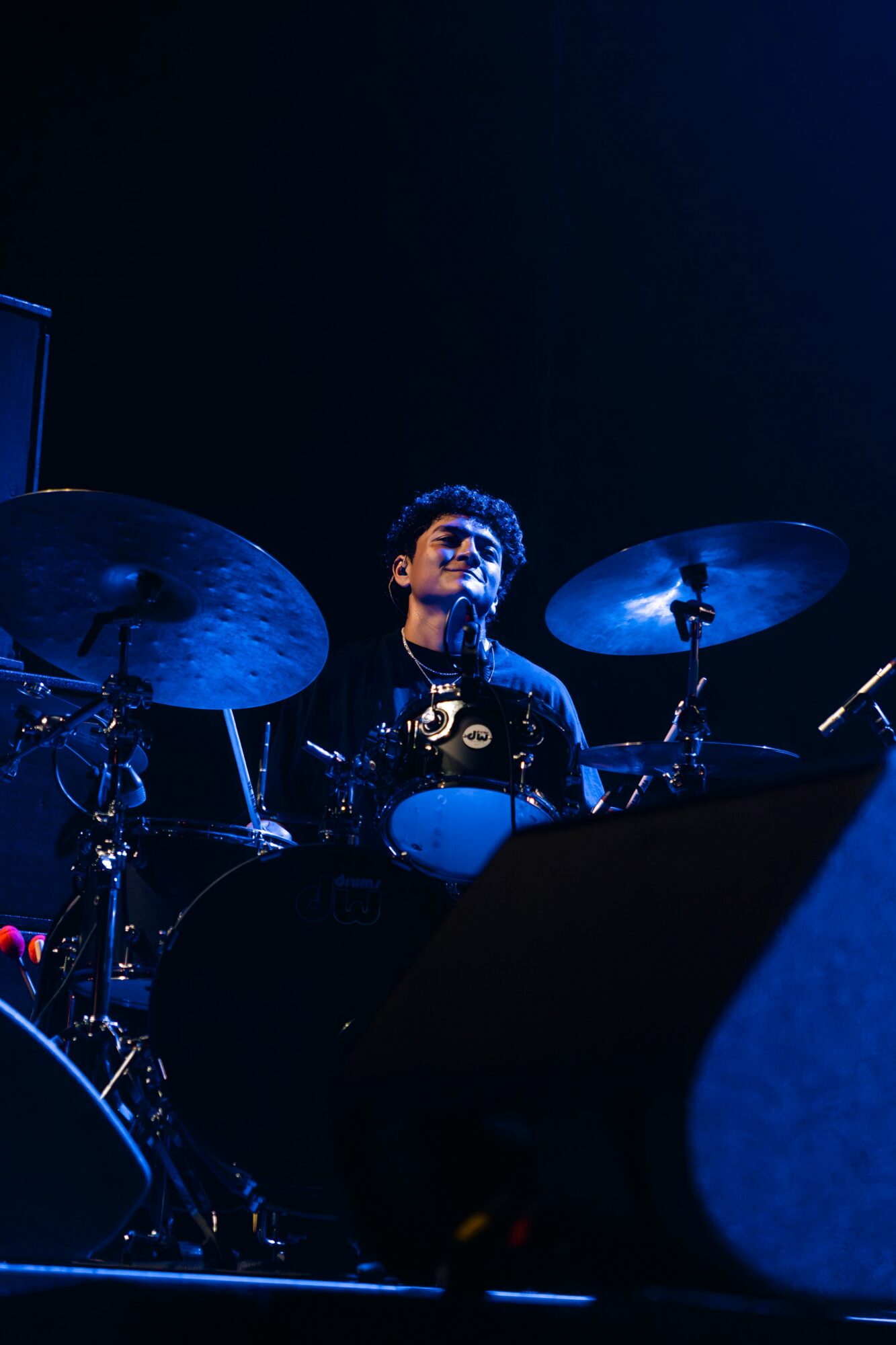 A drummer playing on stage with blue lighting, surrounded by drums and cymbals, focused on performance.