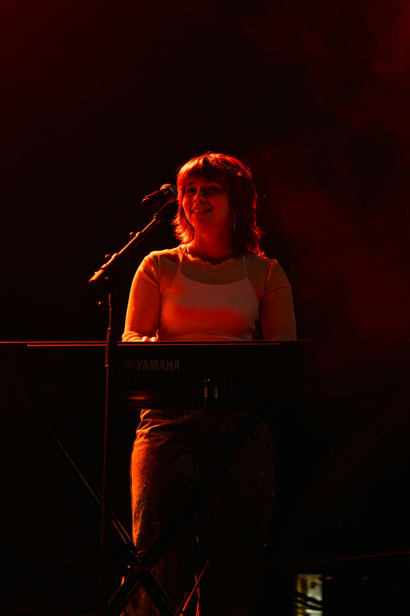 Person playing keyboard on stage with dark background, illuminated by red light, facing forward.
