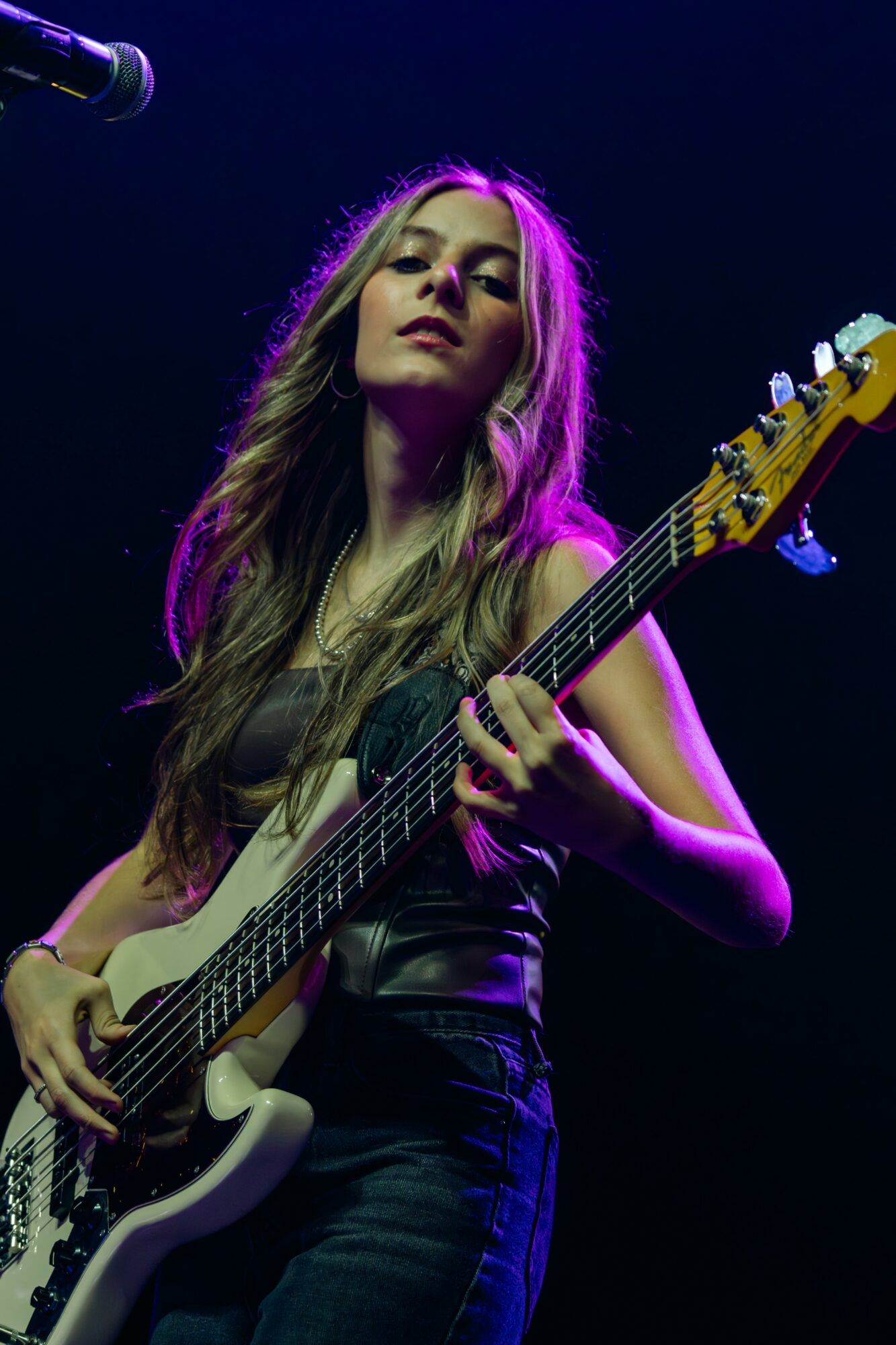 Female musician playing an electric guitar on stage with purple lighting, looking confident, long hair, dark outfit.