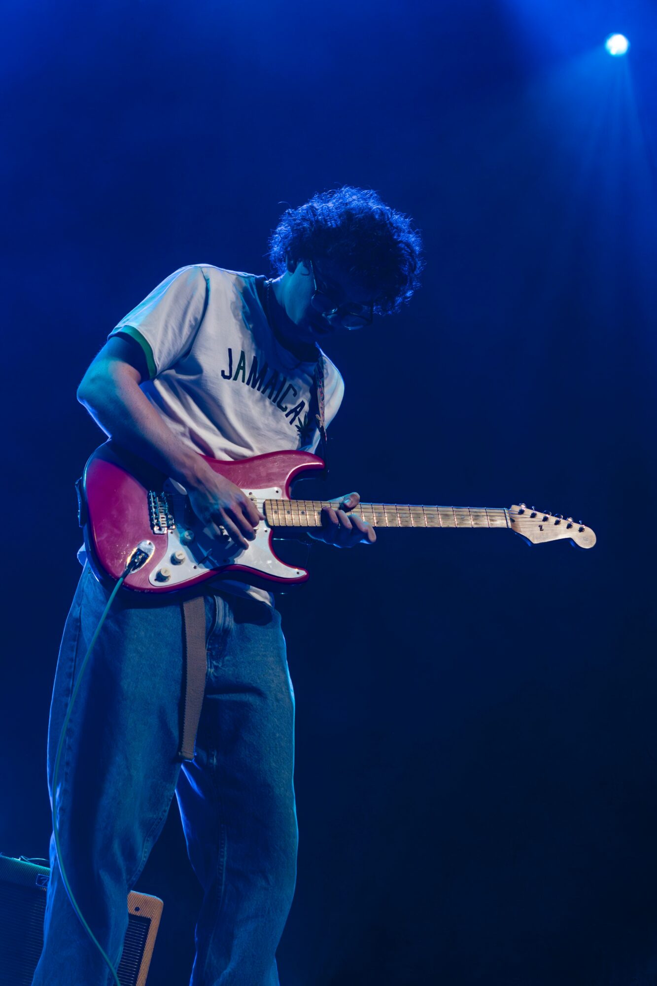 Person playing a red electric guitar on stage with blue lighting, curly hair, wearing a white and green shirt and blue pants.