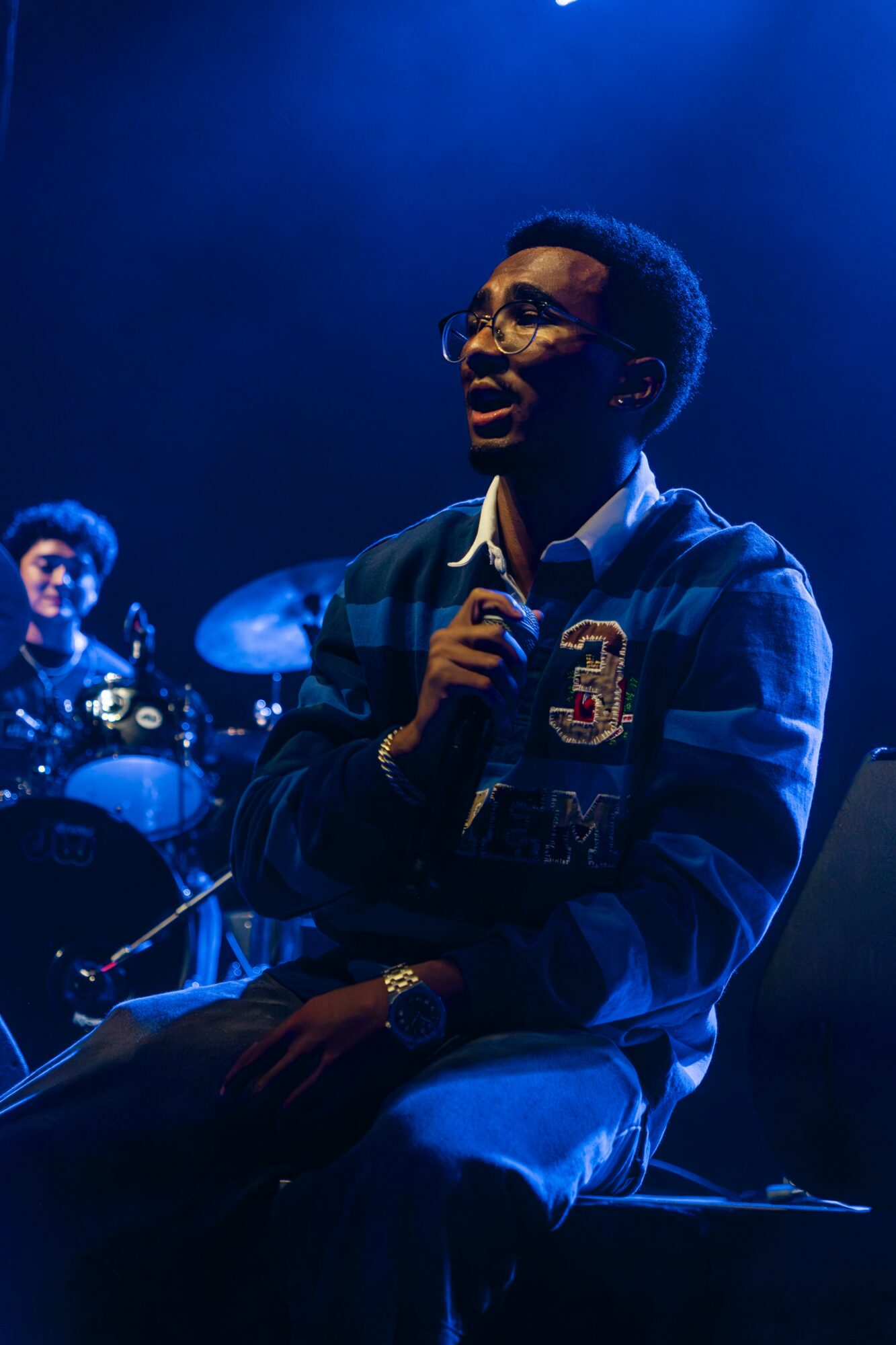 Young man with glasses singing into a microphone on stage, with a drummer in the background, under blue lighting.