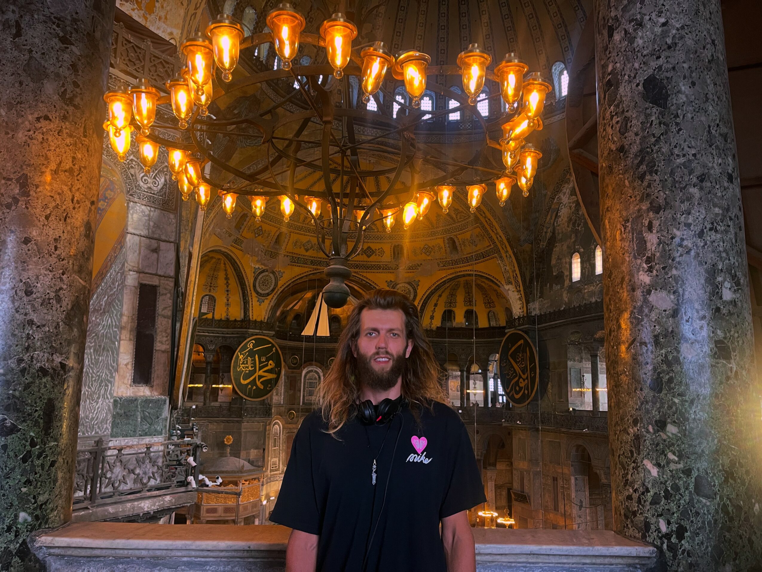 Person with long hair and beard standing inside a building with a large chandelier overhead, ornate architecture in background.
