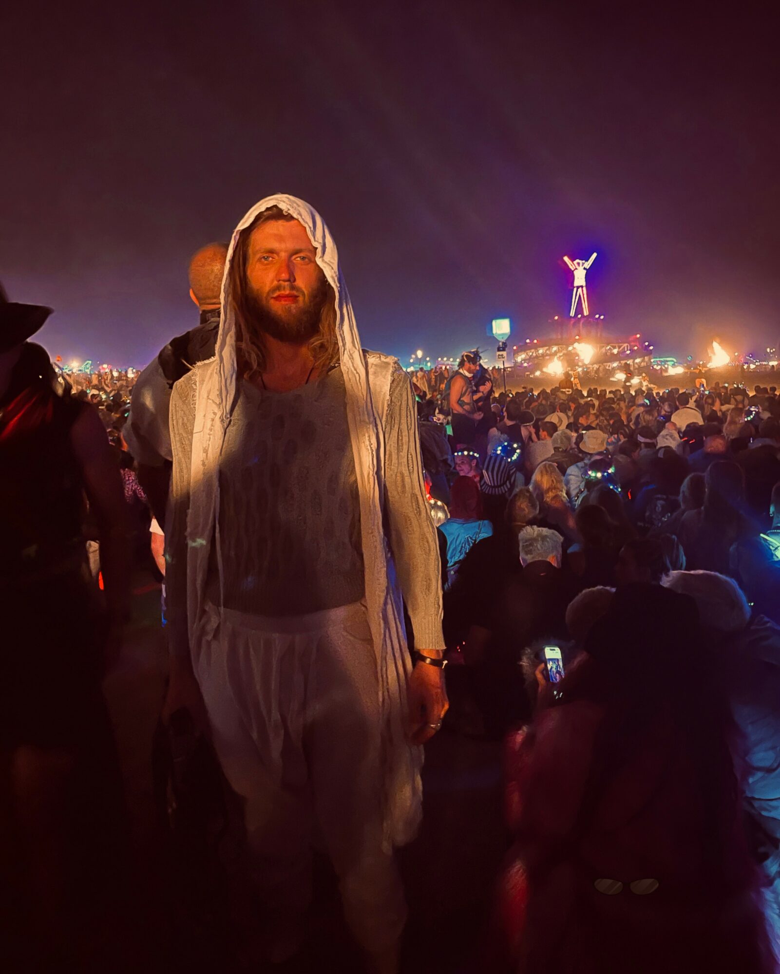 Person with long hair and beard wearing a hooded jacket at night festival, crowd and a lit figure in the background.