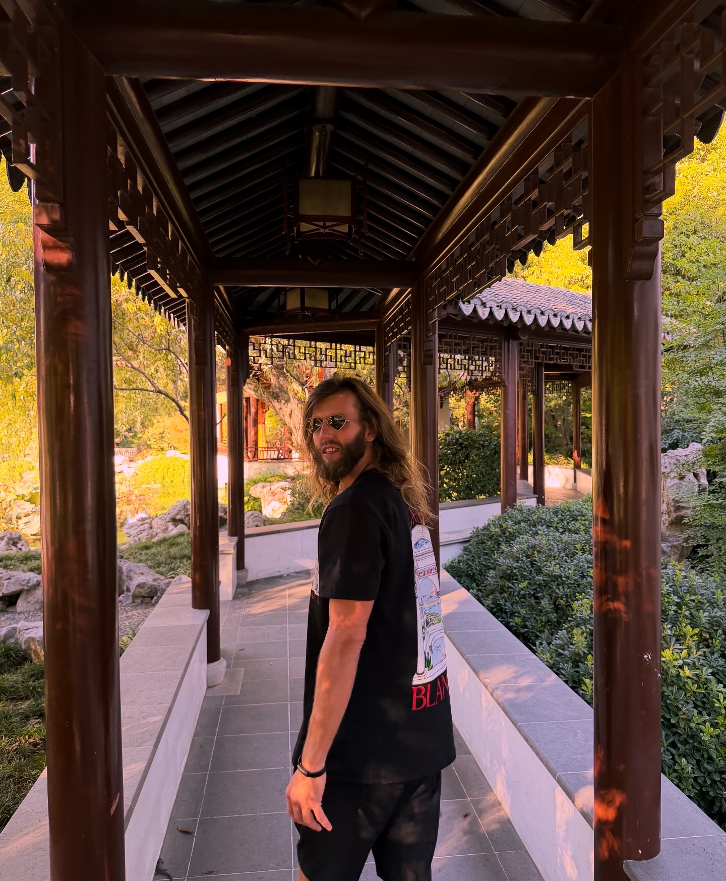 Person with long hair and glasses standing in a traditional wooden corridor outdoors, surrounded by greenery.