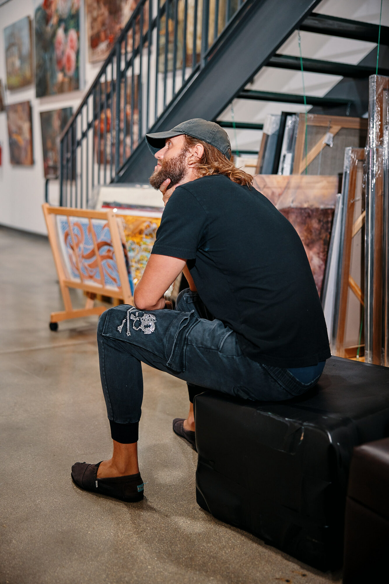 Person with long hair and beard sitting on a black ottoman, wearing a black t-shirt, jeans, and a cap, in an art gallery.