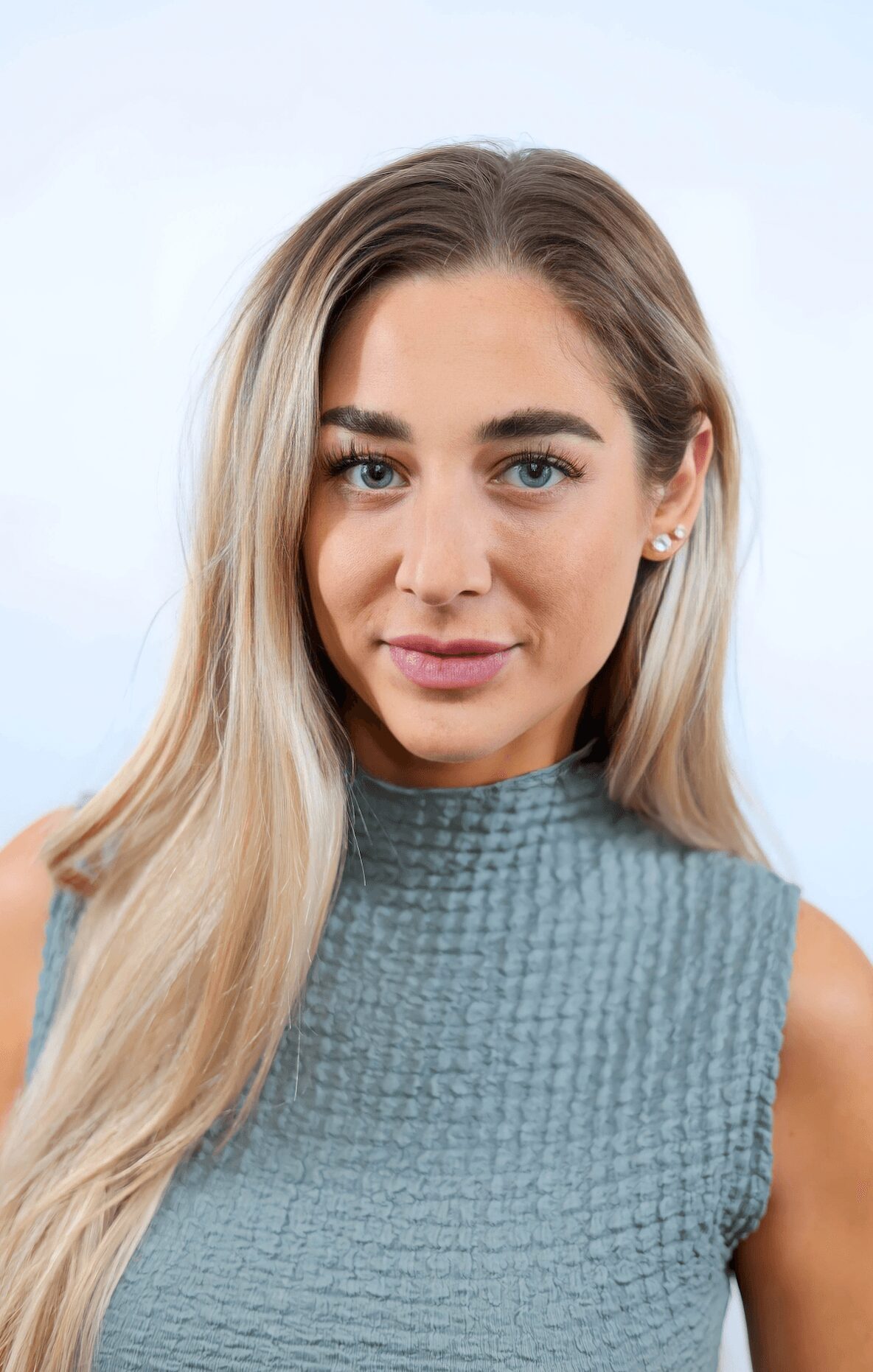 Young woman with long blonde hair and blue eyes, wearing a sleeveless textured blue top, smiling slightly, against a light background.