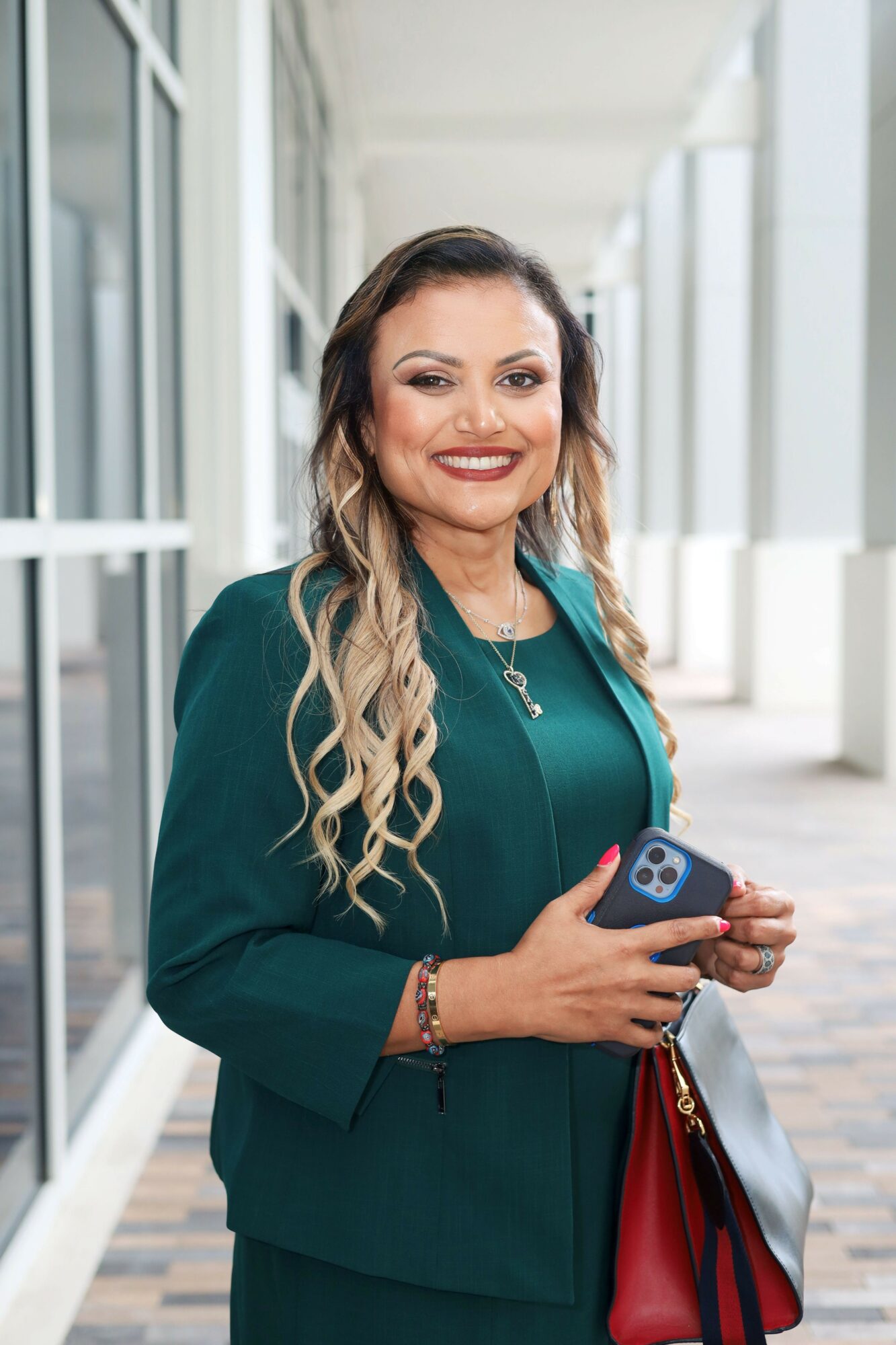 Woman with long wavy hair in a teal blazer holding a smartphone, smiling, in a modern building corridor.
