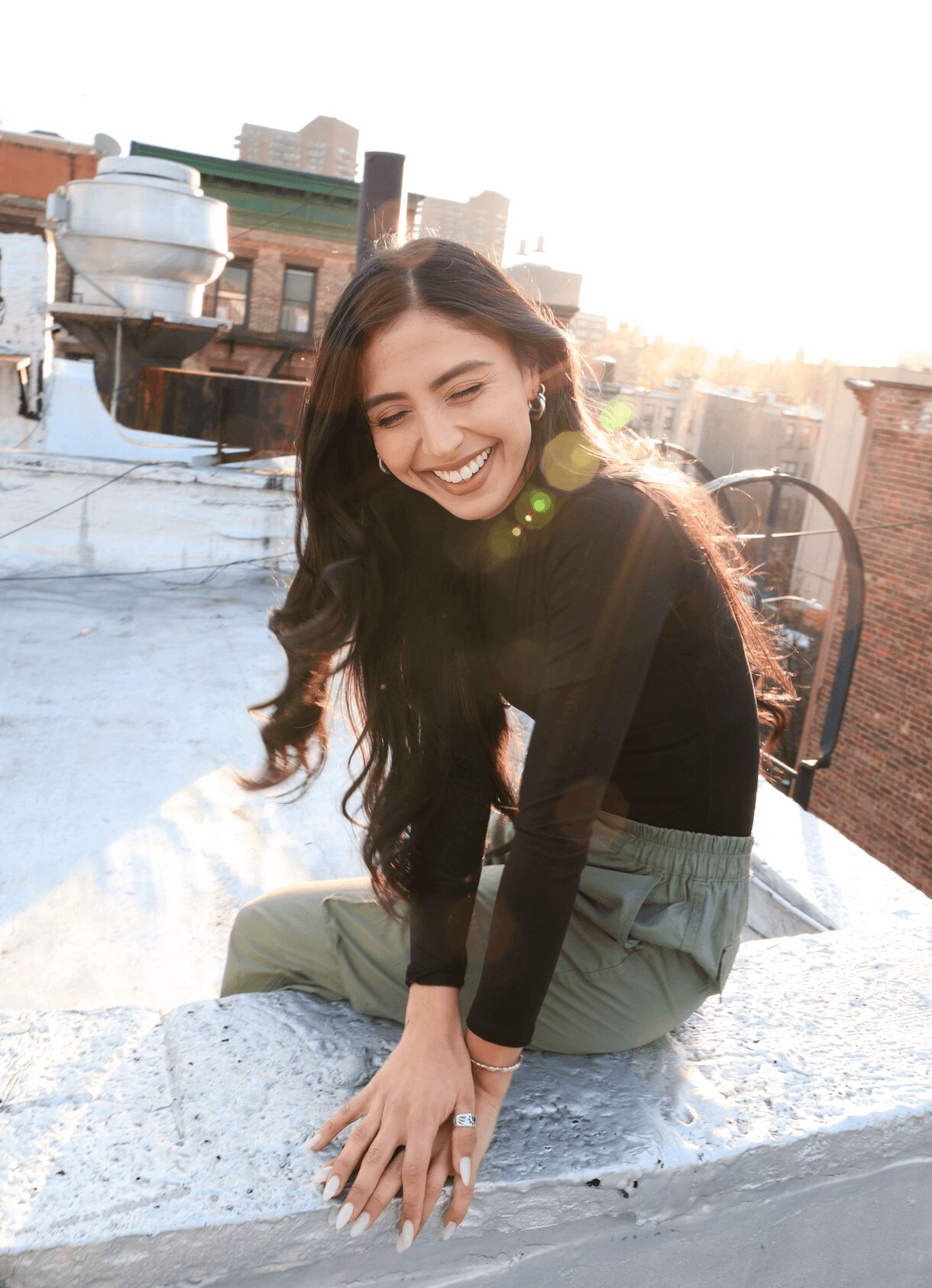 Young woman with long dark hair smiling, sitting on a rooftop ledge with city buildings in background, sunlight shining.