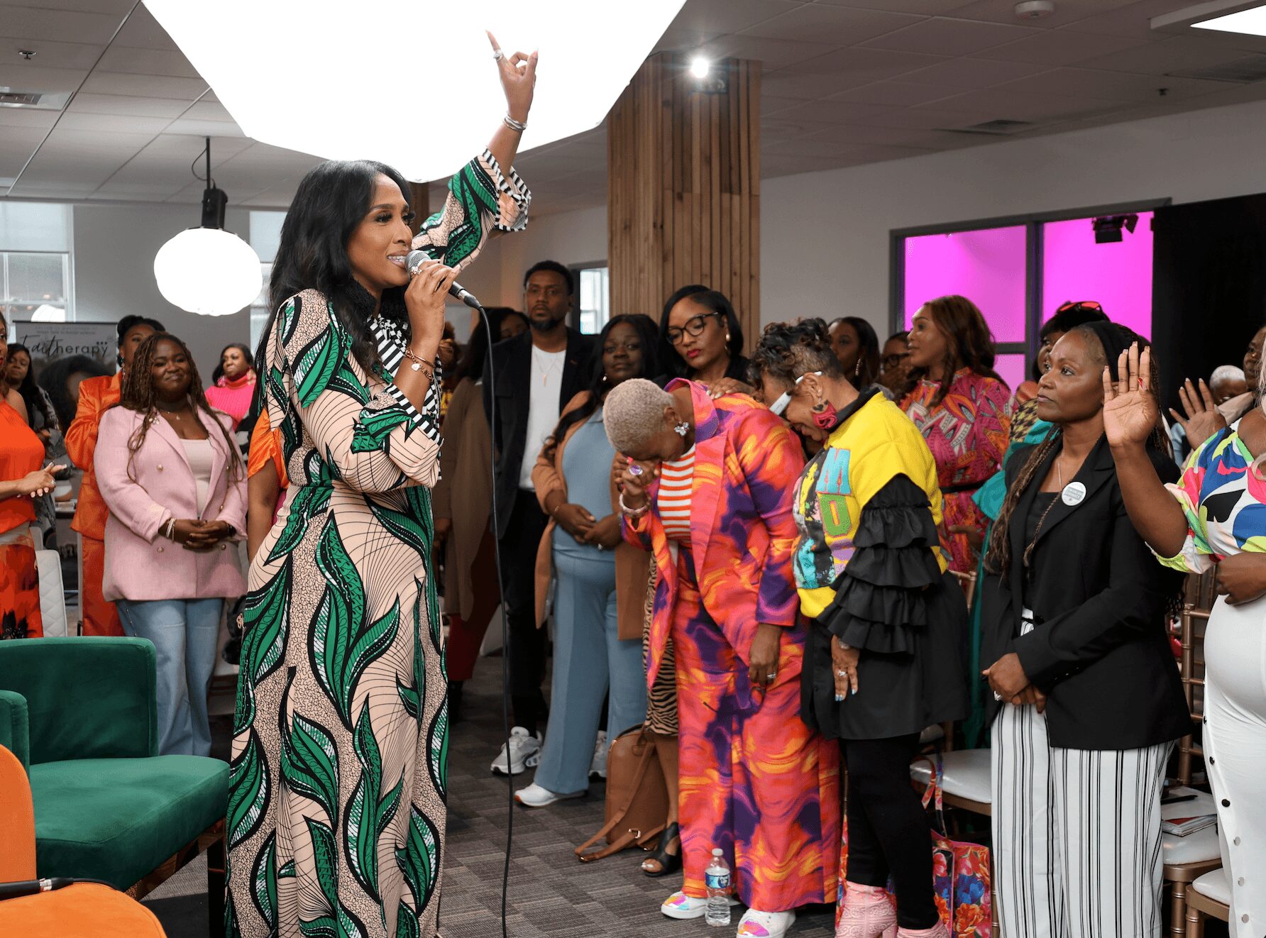 Woman in a patterned dress speaking into a microphone while a group of people listen attentively in a modern indoor space.
