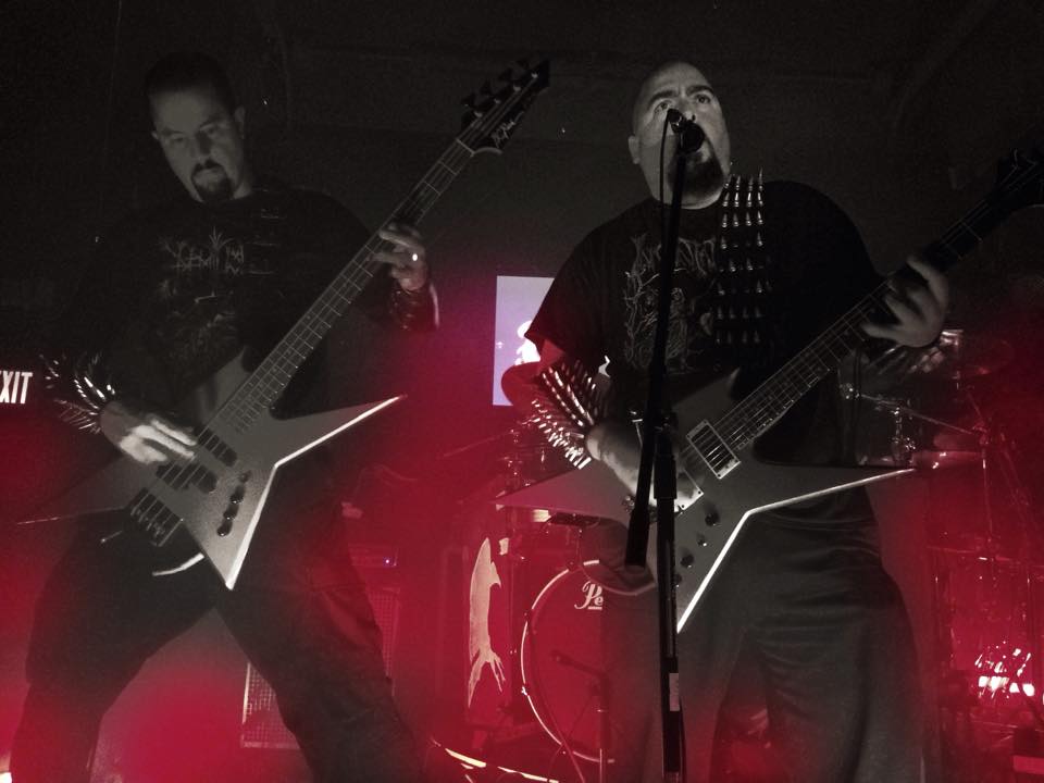 Two musicians perform on stage with guitars, one singing into a microphone, red stage lighting, and a drum set in the background.