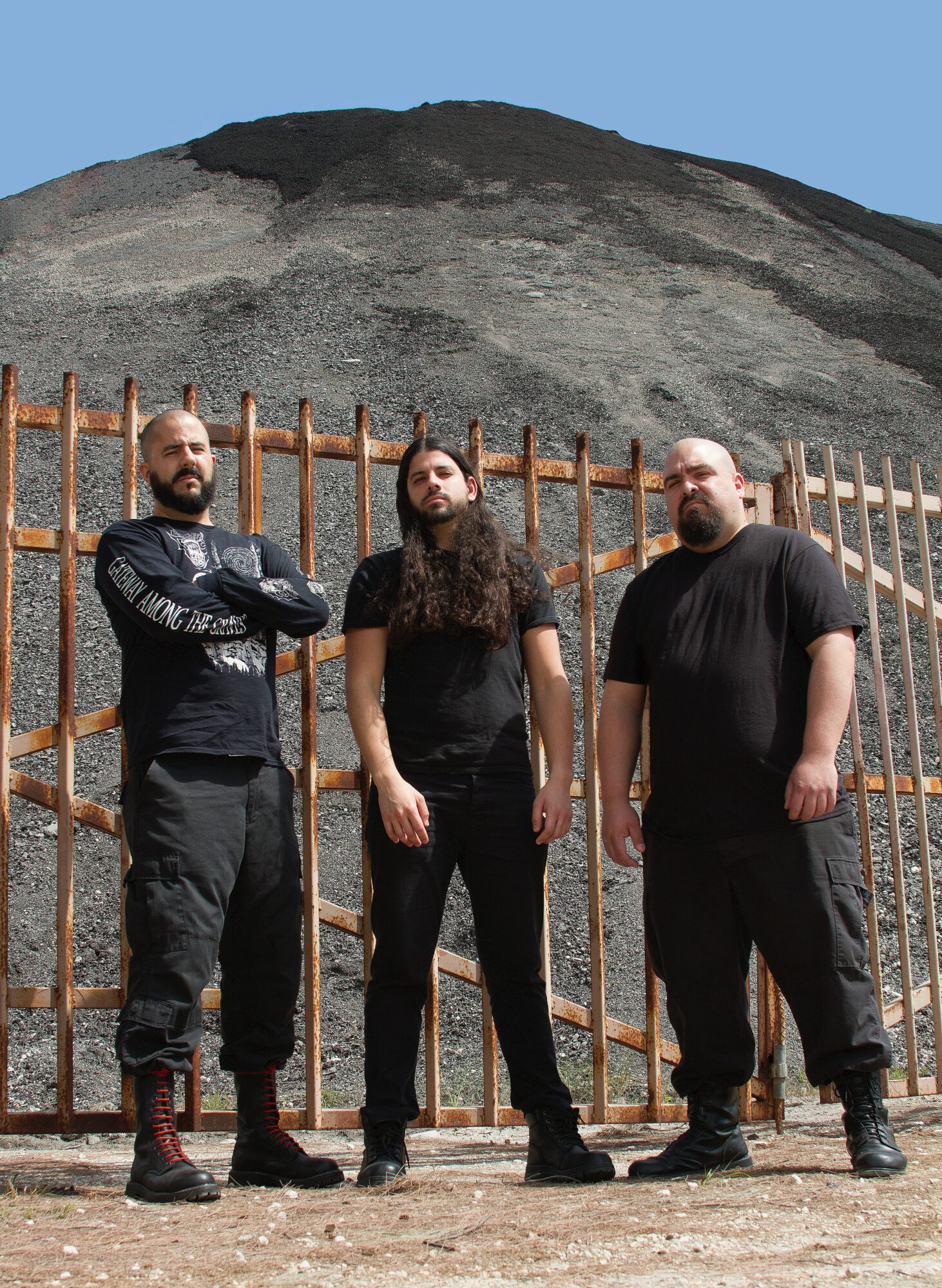 Three men stand in front of a metal fence with a mountain in the background, under a clear blue sky.