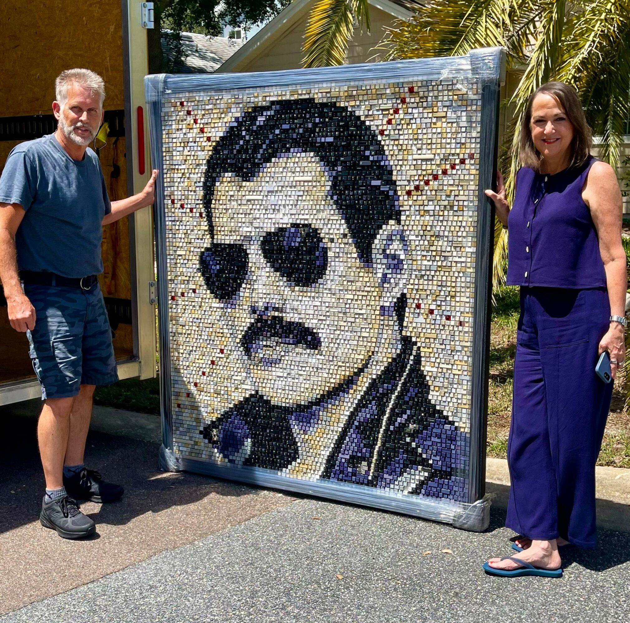 Two people stand beside a mosaic portrait of a man with sunglasses and a mustache, outdoors with trees and a building behind.