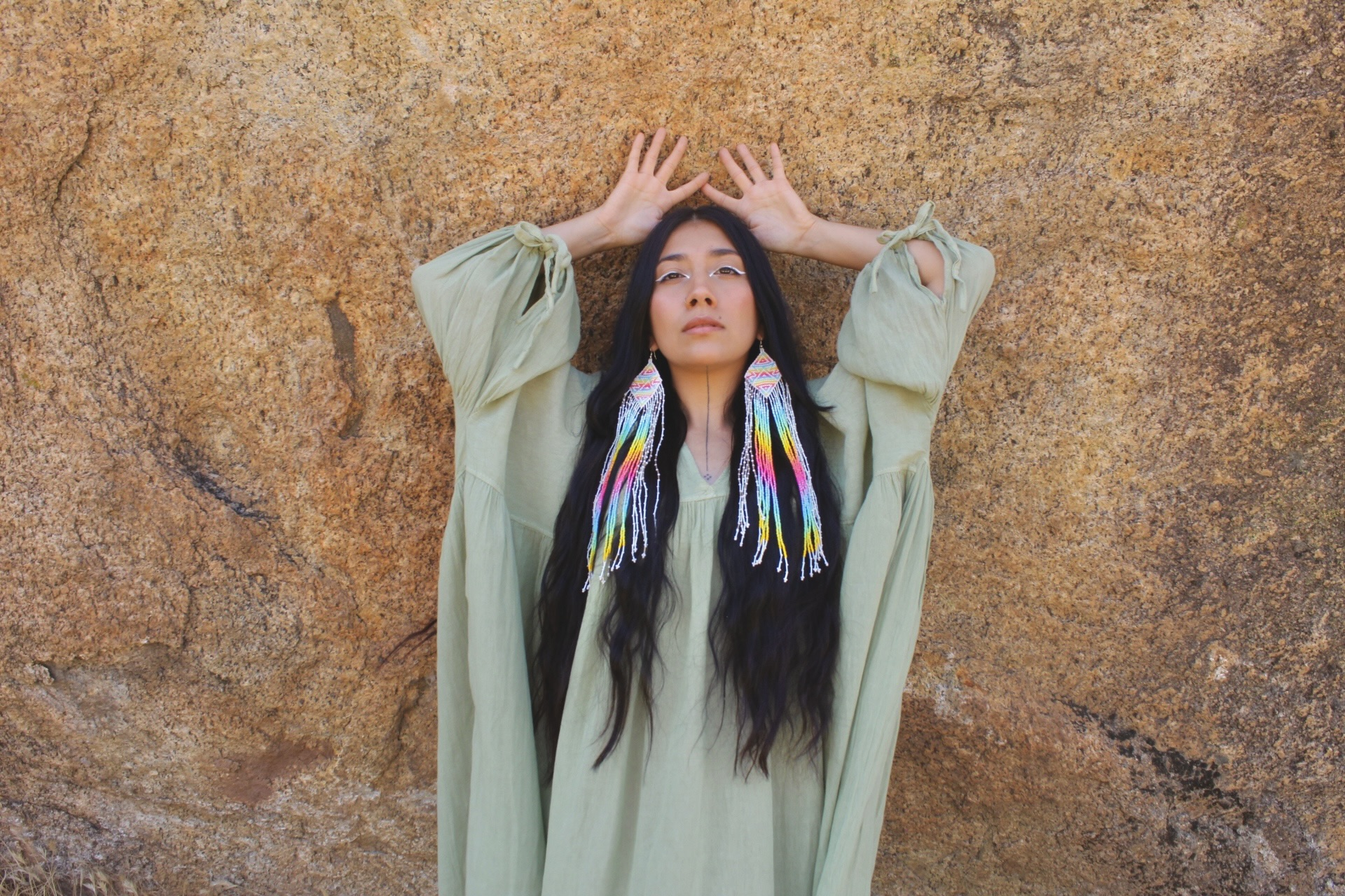 Woman with long dark hair and colorful scarf, standing against a textured wall, hands raised above her head.